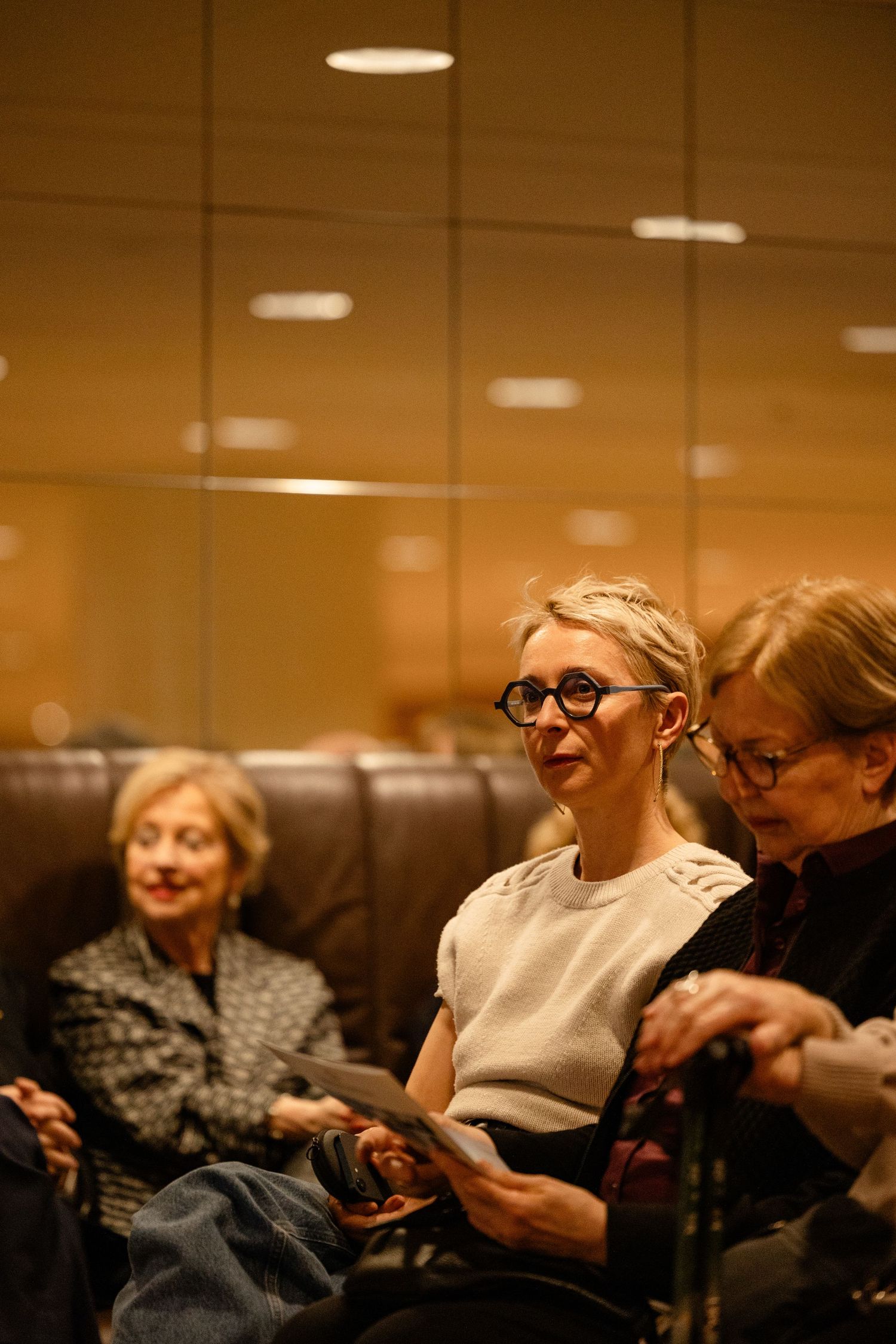 Three women seated in a warmly lit auditorium, middle woman wearing bold black glasses and white top, attentively watching an event.