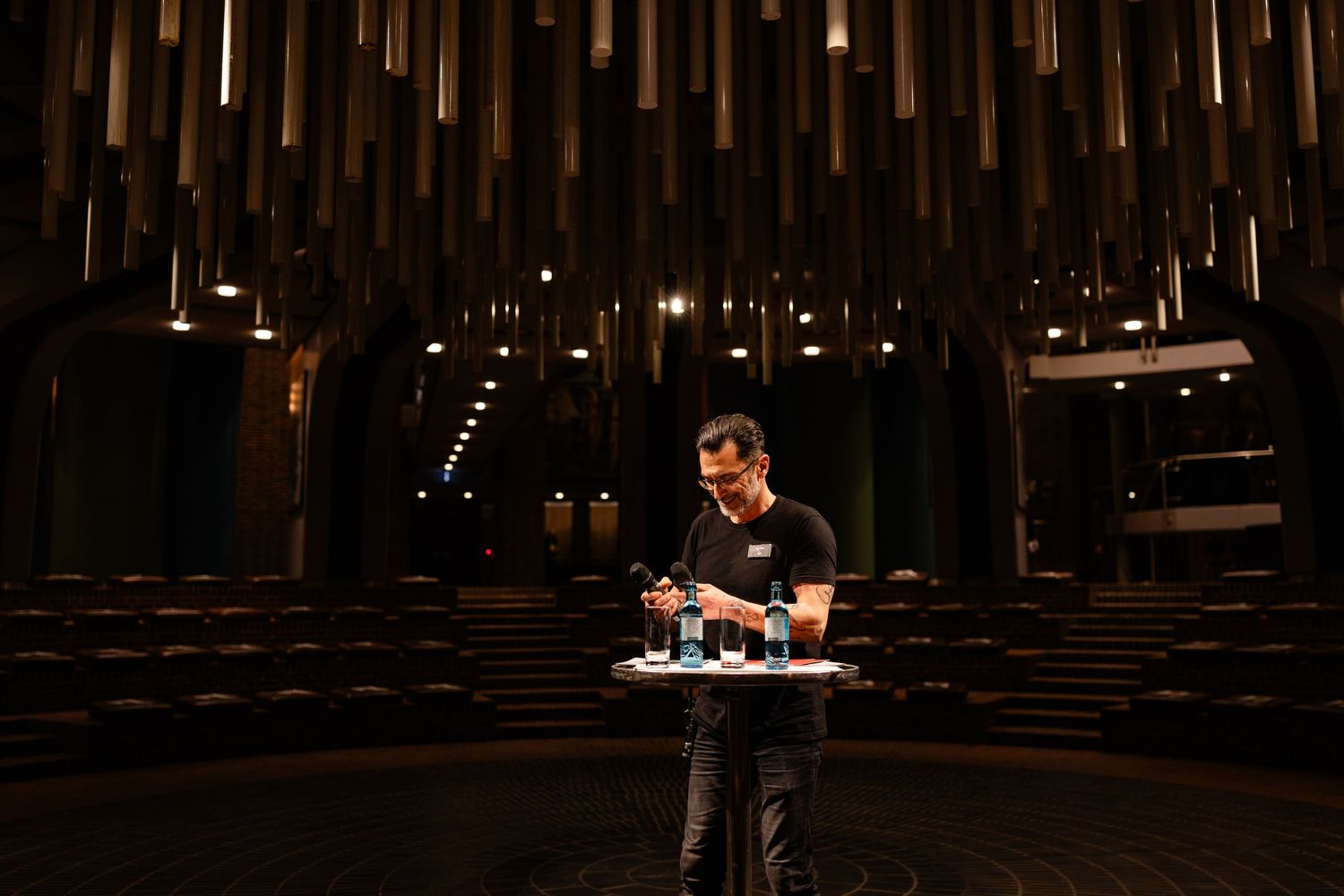 Man standing at a podium in a dramatic modern auditorium with wooden ceiling installations and rows of empty seats.