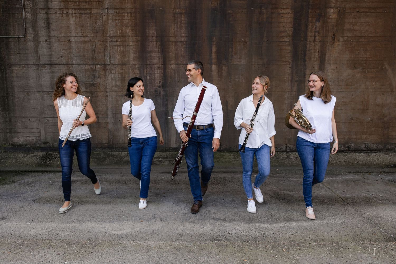 Five musicians in white shirts and jeans walking and holding instruments including flute, clarinet, saxophone, and French horn.
