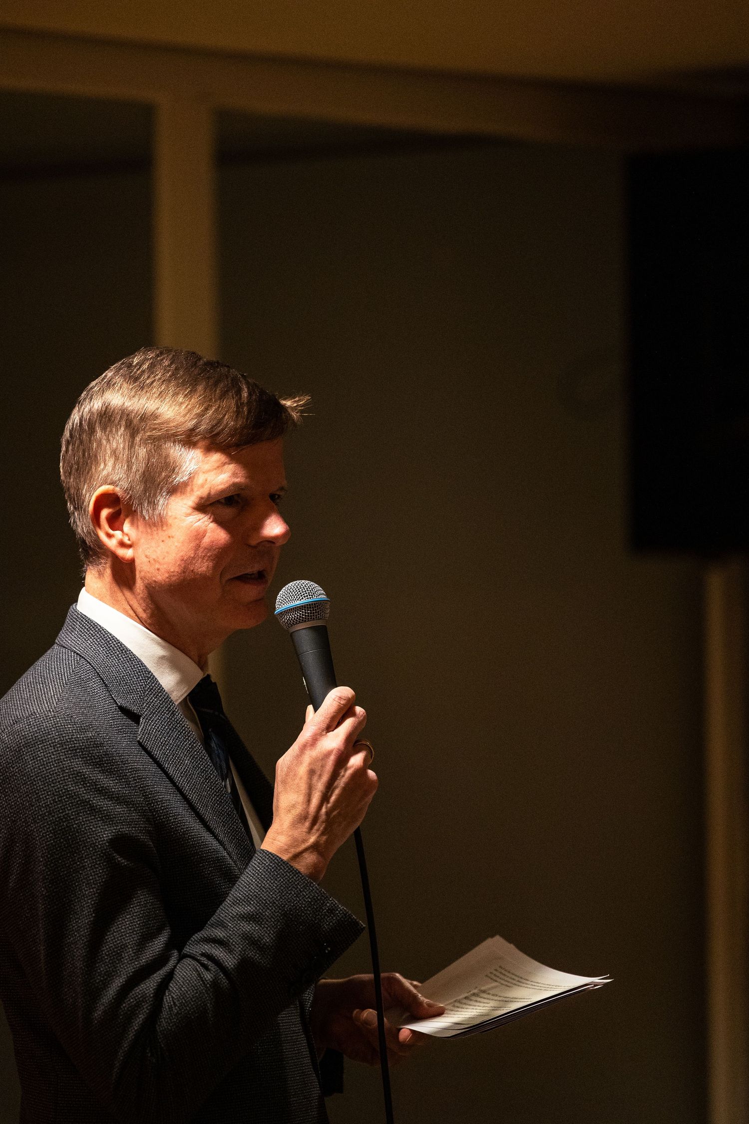 Man in suit speaking into microphone while holding papers at a formal event or conference.