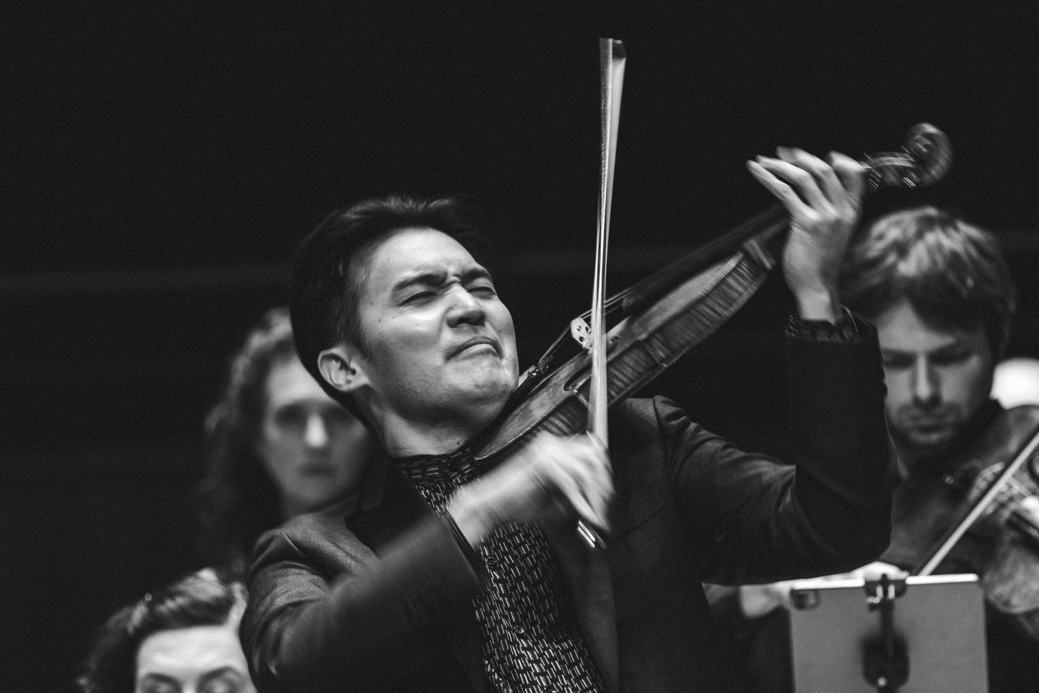 Black and white photo of a focused male violinist performing with an orchestra in the background.