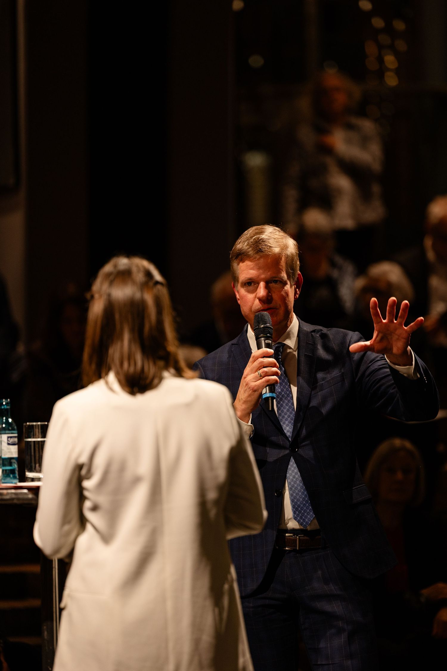 Man in blue suit speaks into microphone while gesturing with hand at formal indoor event.