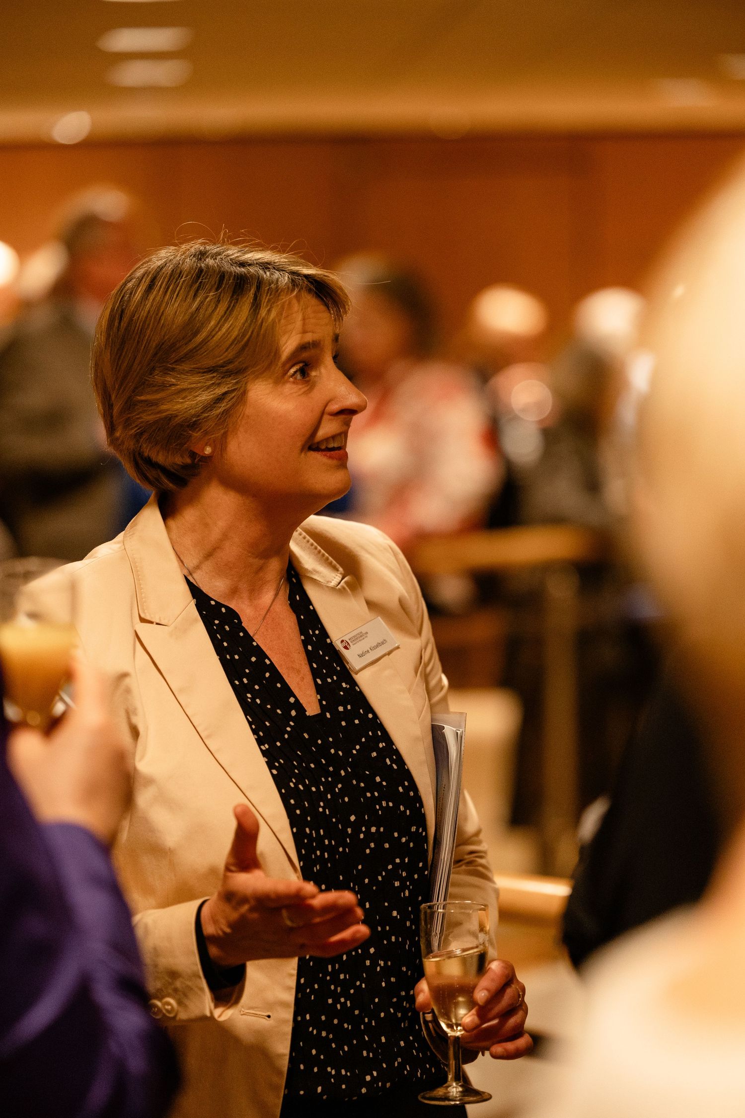 Woman in beige blazer holding champagne glass, engaged in conversation at a formal networking event.