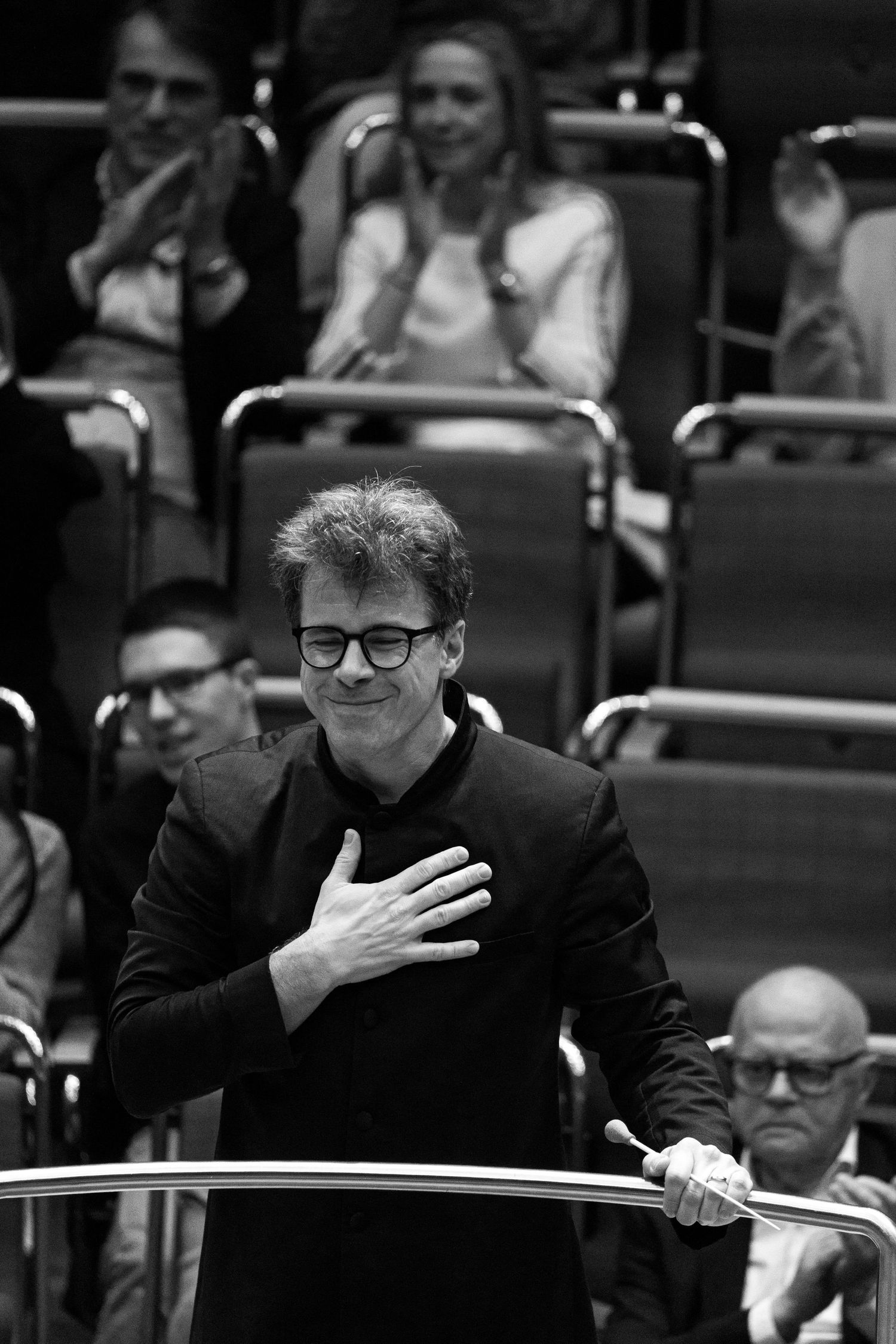 Black and white photo of a conductor holding a baton, placing hand on chest in gratitude before an audience.
