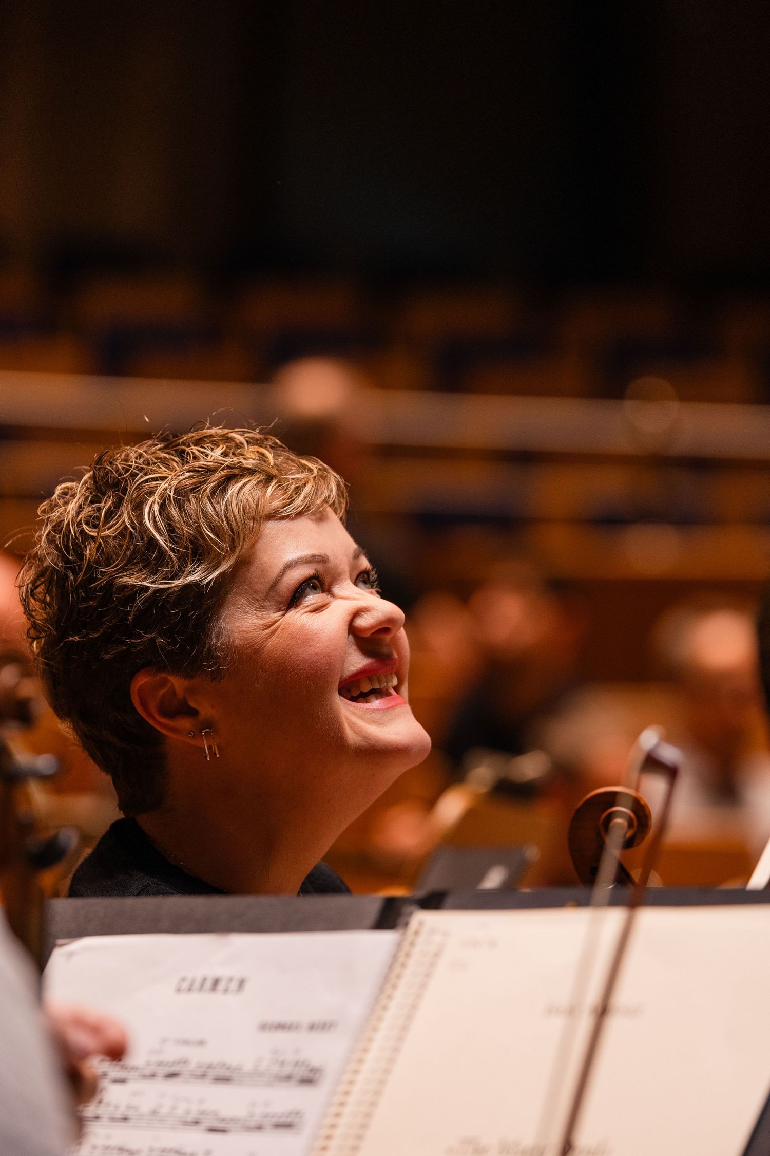 Smiling female musician looks up during orchestra rehearsal with Carmen sheet music on stand in concert hall.