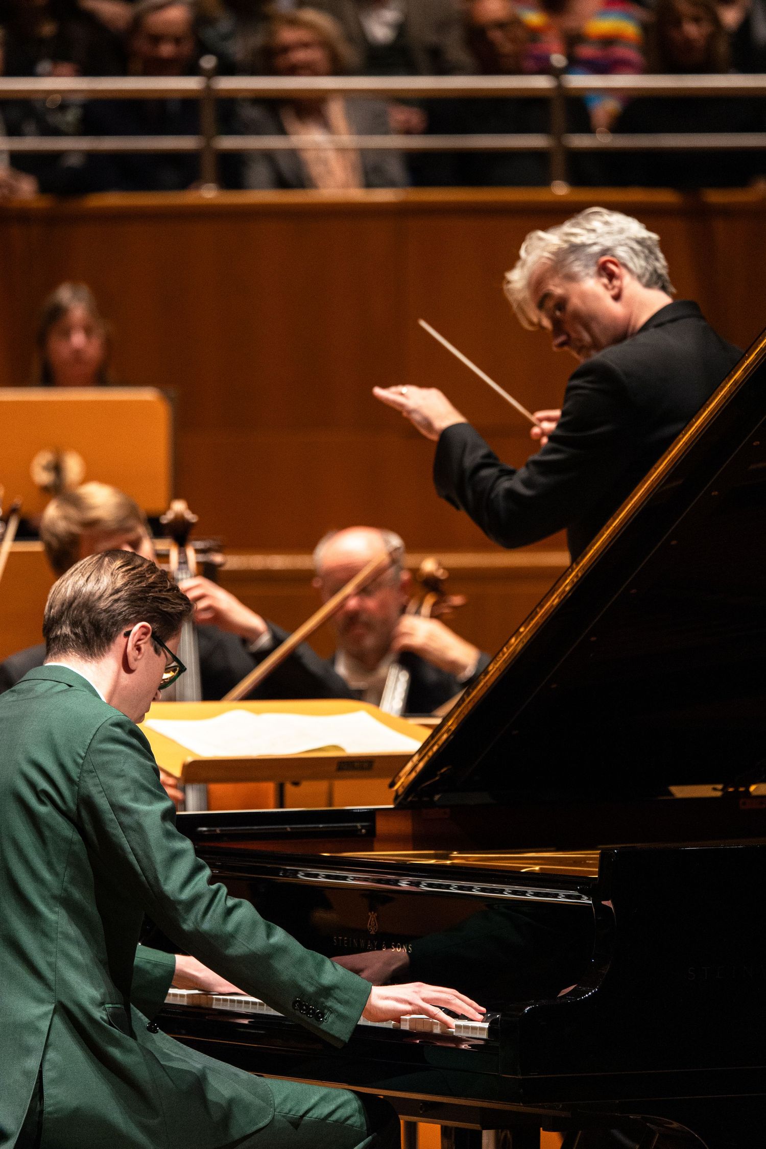 Pianist in green suit performs with orchestra conductor holding baton at grand piano concert hall.