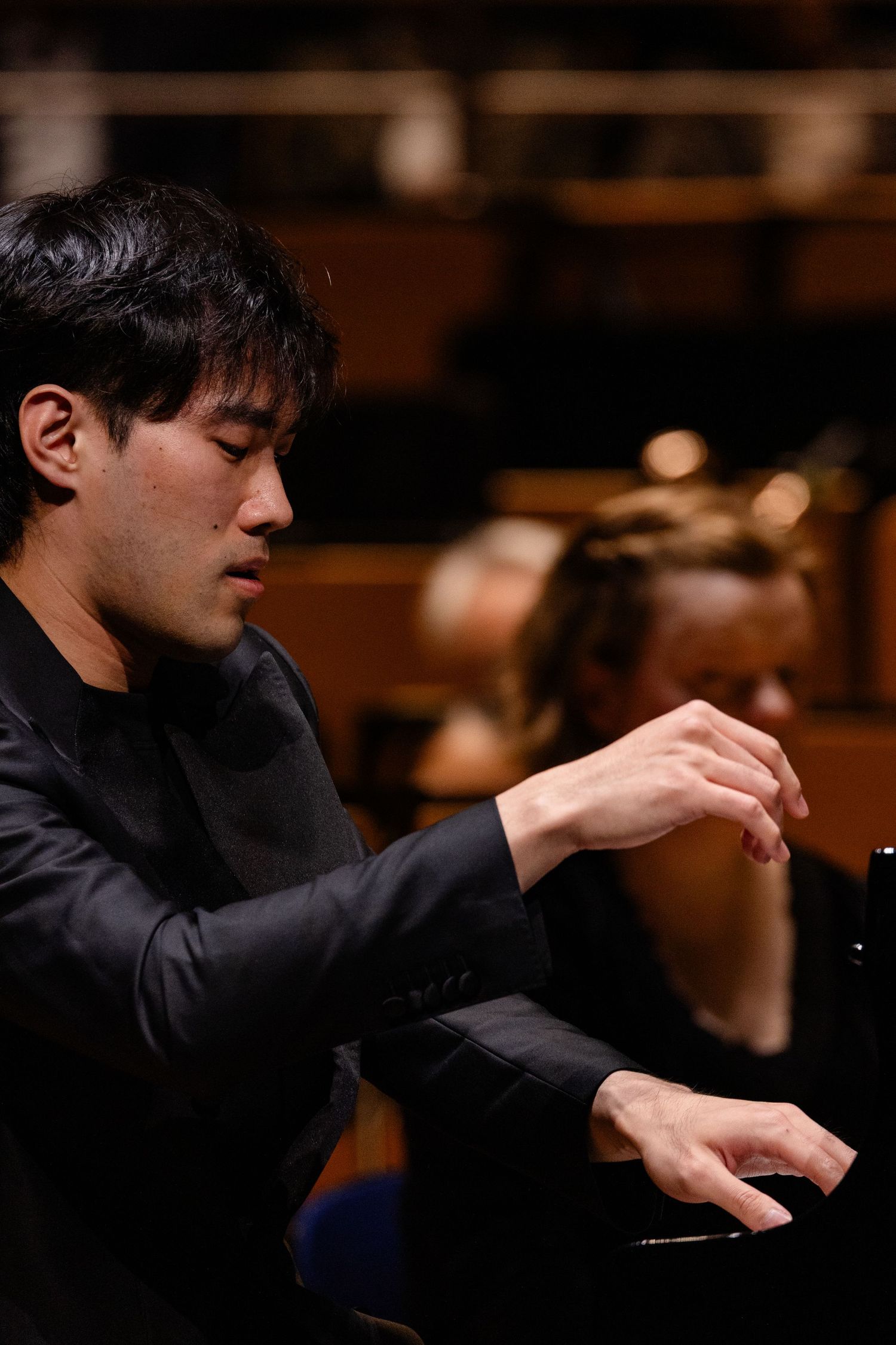 A focused male pianist performs on stage in a black jacket, hands poised over keys in a concert hall.