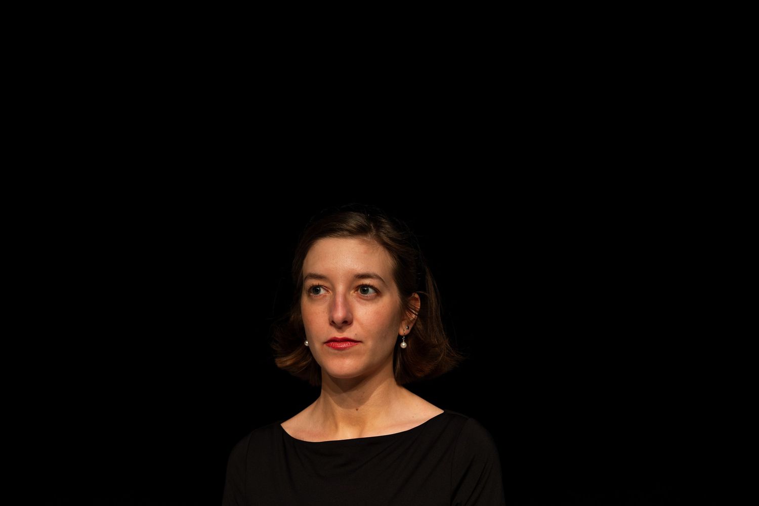 Woman with short brown hair wearing a black top, posed against a dark background in a professional portrait.