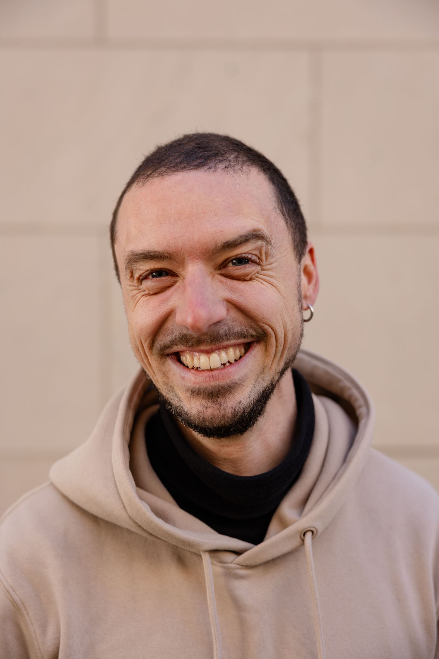 Smiling man with short hair, ear piercing, and beard wearing a beige hoodie over black shirt against a light wall.