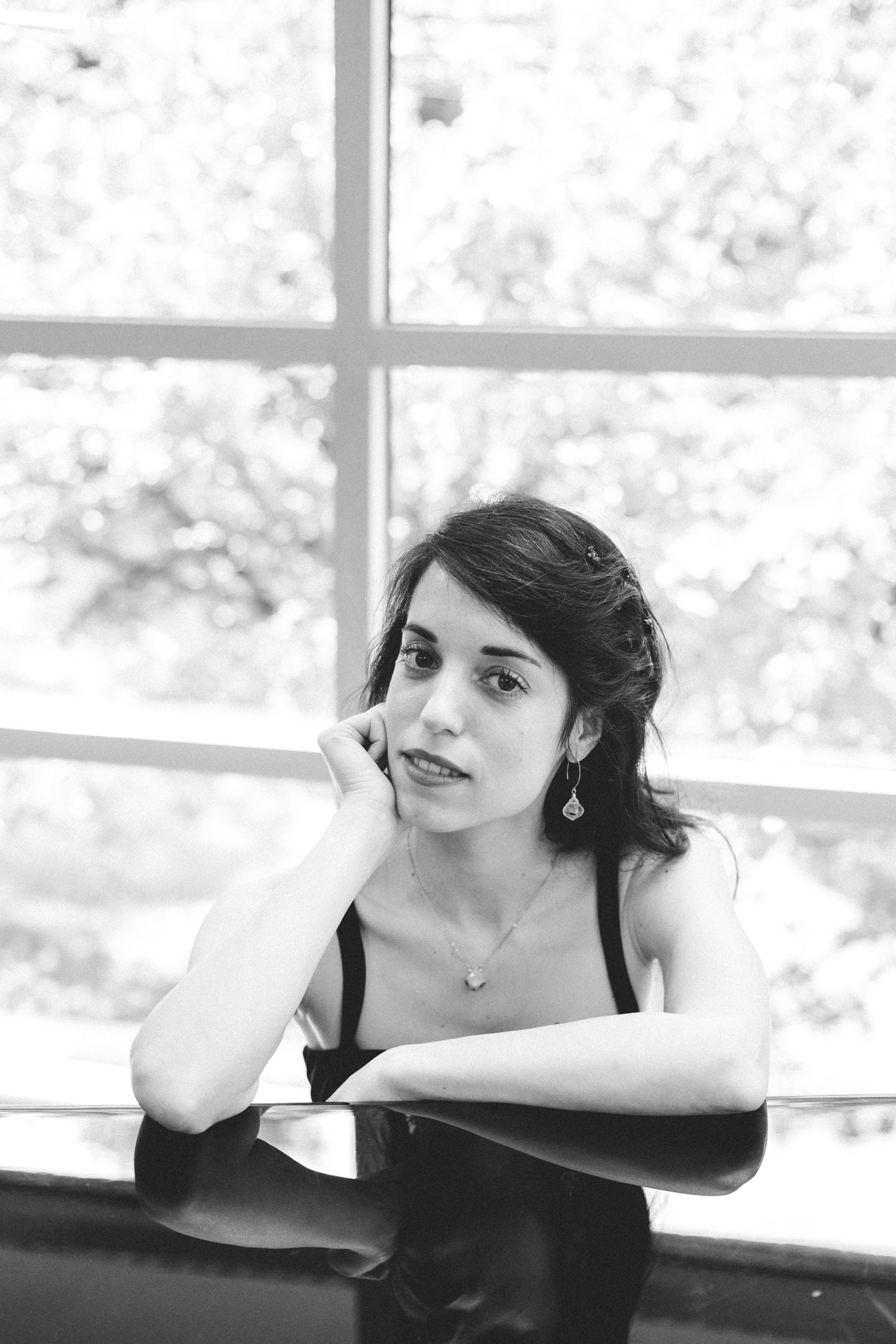Black and white portrait of a young woman resting her chin on her hand near a large window with soft natural light.