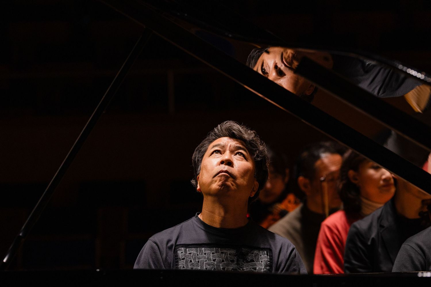 A musician gazes upward beside a grand piano in a dimly lit concert hall setting.