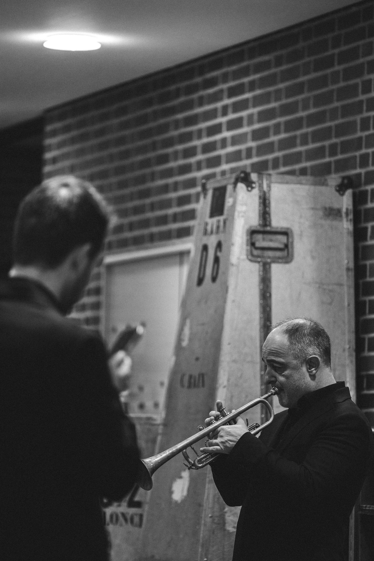Black and white photo of a man playing trumpet near a brick wall while another man watches nearby.