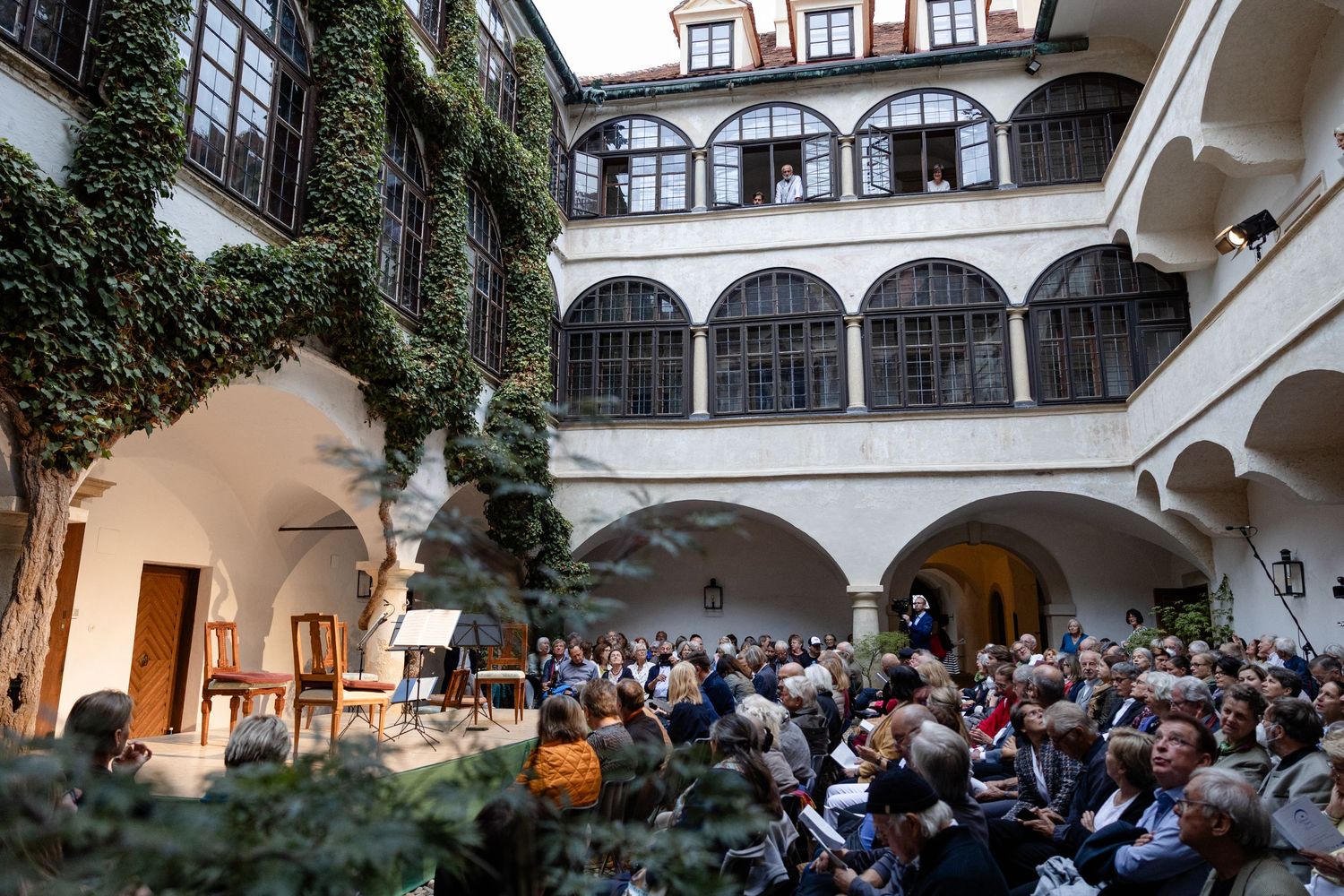 Crowd gathered in a historic courtyard with arched arcades and ivy-covered walls for an outdoor cultural event.