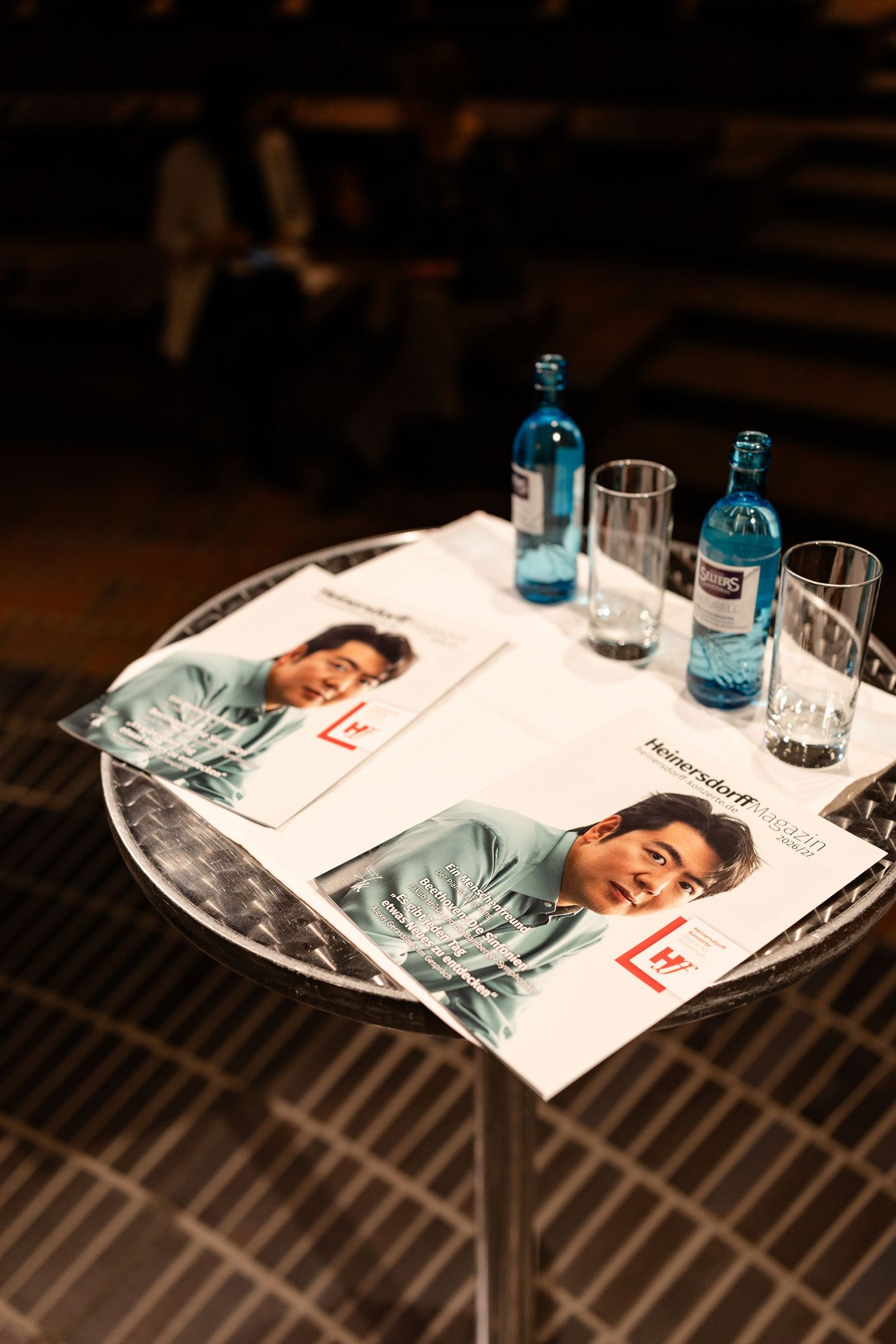 A round table with magazines featuring a man on the cover, blue water bottles, and empty glasses at an event.