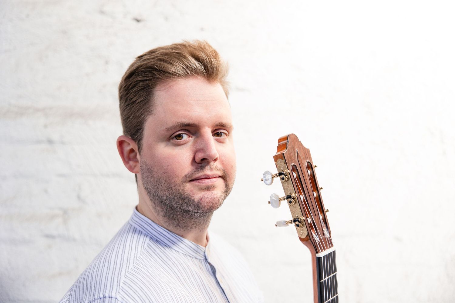 Young man holding a classical acoustic guitar against a white brick wall, looking over his shoulder.