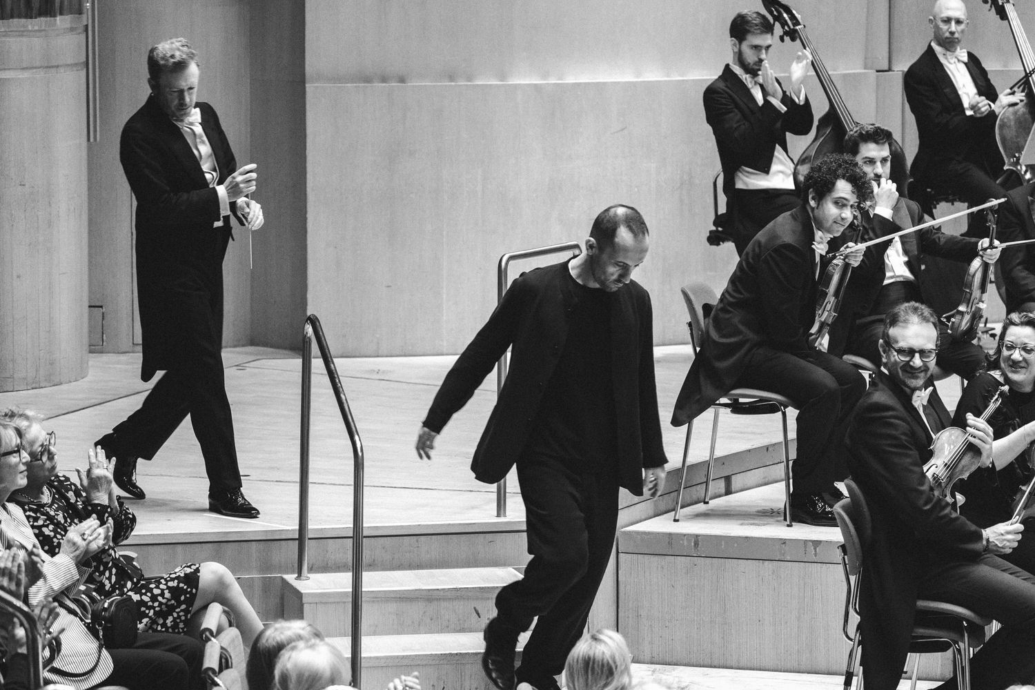 Black and white photo of musicians performing in a concert hall with conductor descending steps.