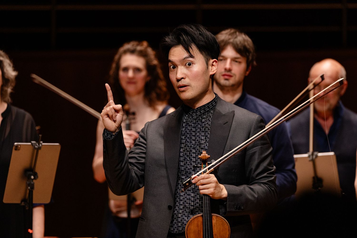 Violinist performing on stage, gesturing with bow in hand, musicians with instruments visible in background.