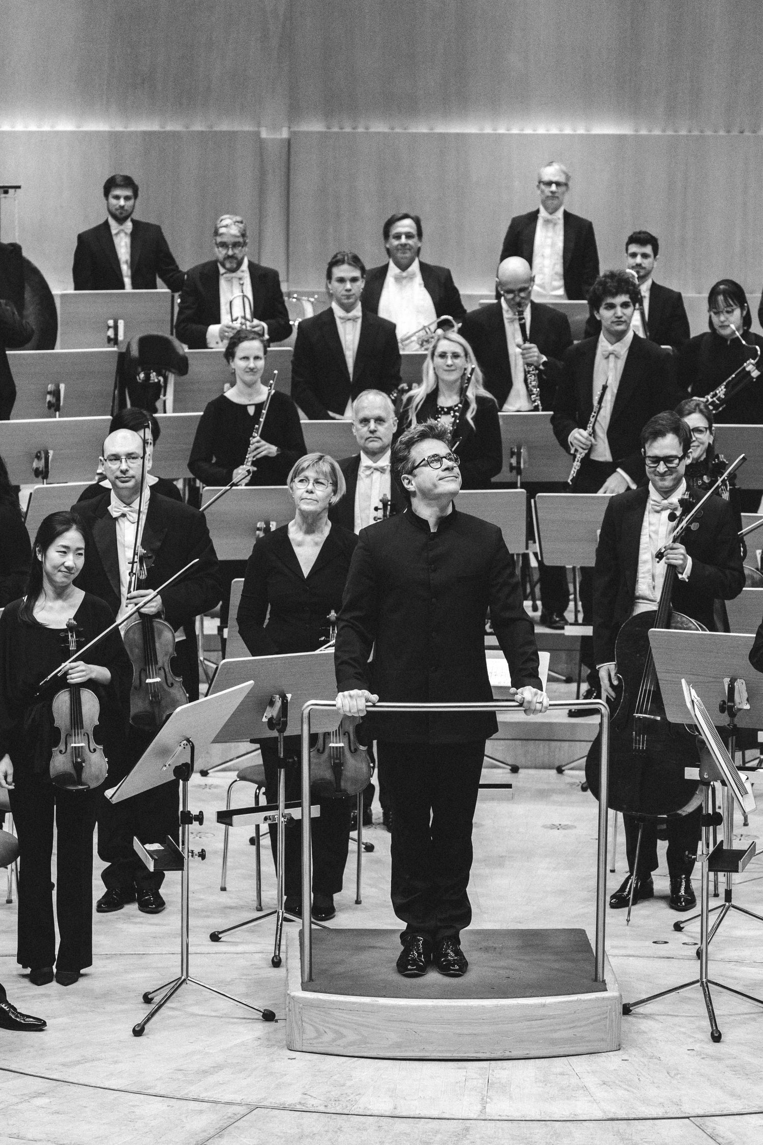 Black and white photo of an orchestra conductor standing at podium with musicians holding instruments behind him.