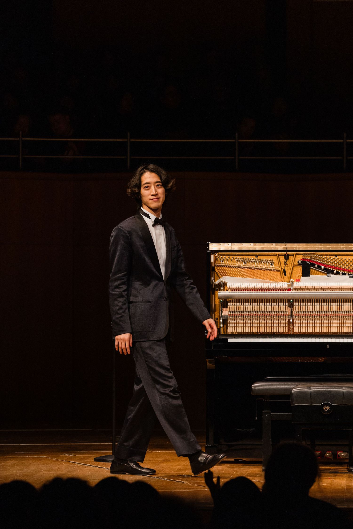 Young male pianist in a tuxedo standing beside an open grand piano on a concert hall stage.