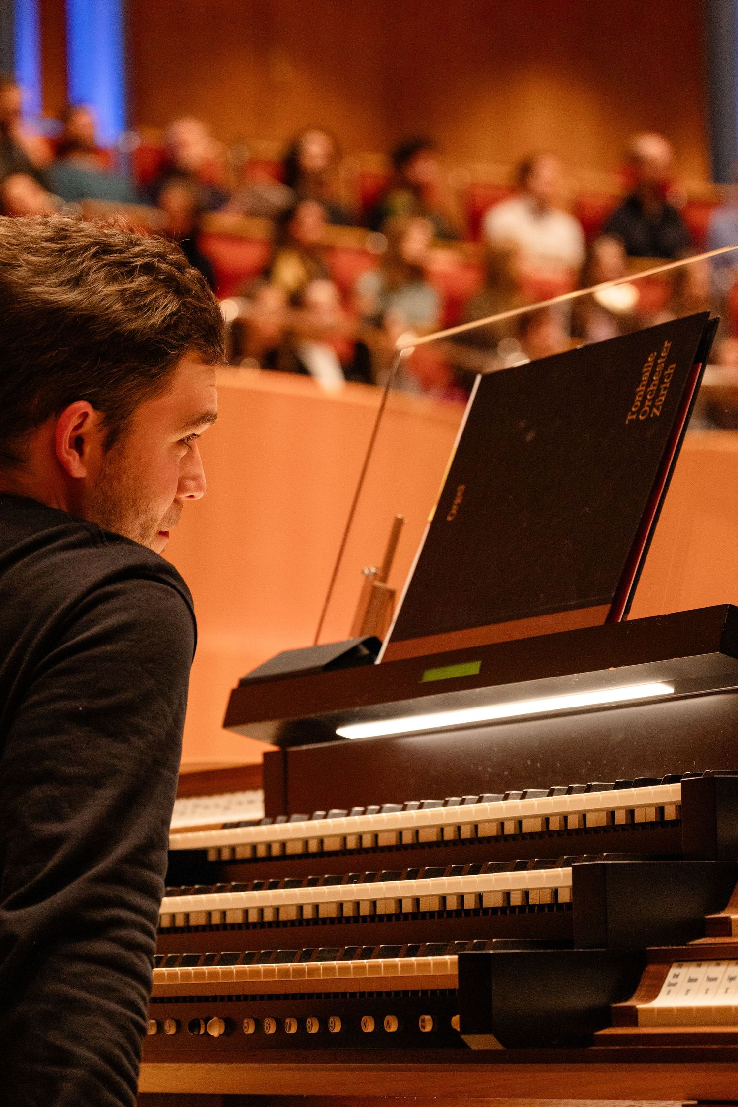 A young man plays a large pipe organ console on stage before an audience in a concert hall.
