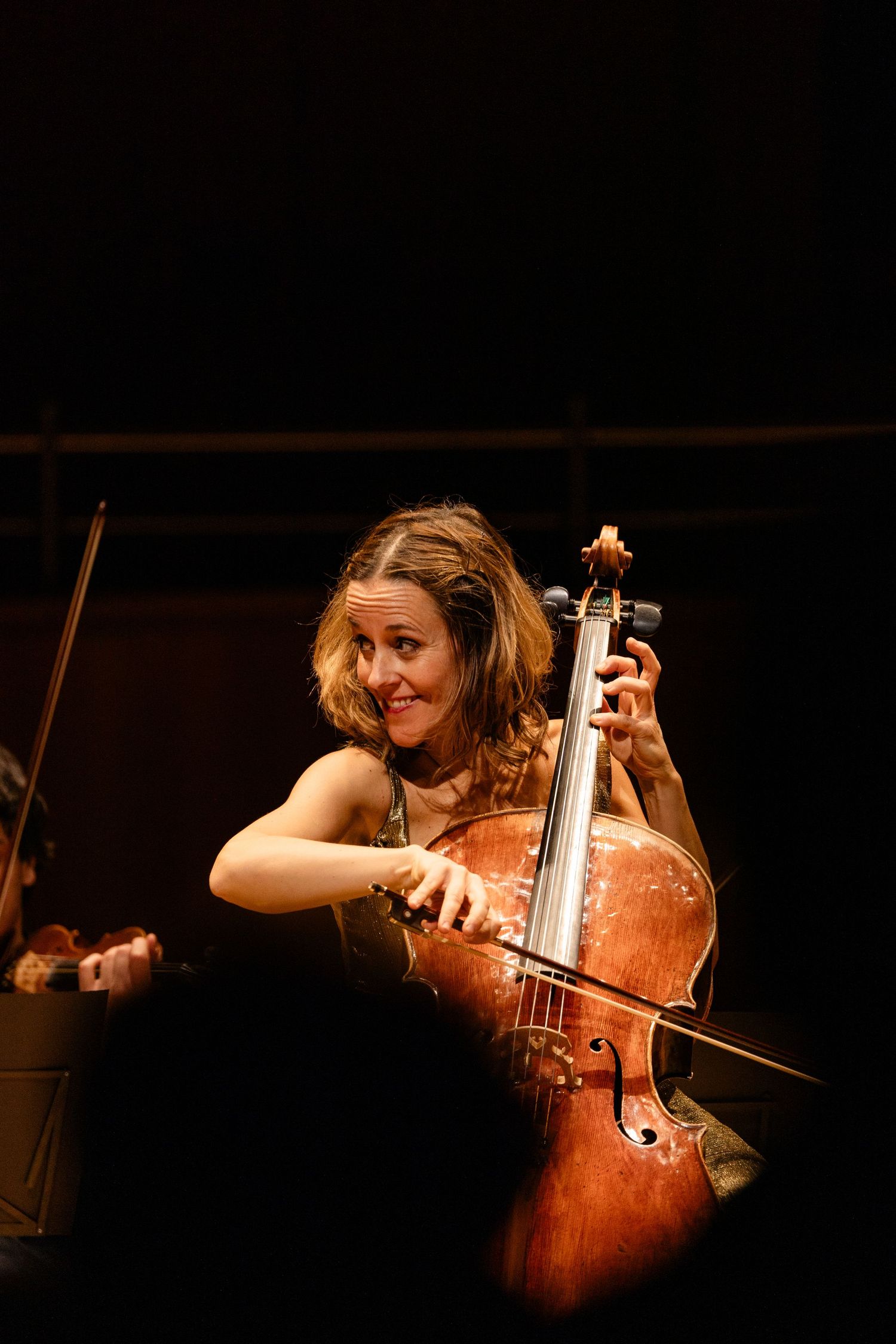 Smiling female cellist performing on stage, playing a wooden cello under dark concert lighting.