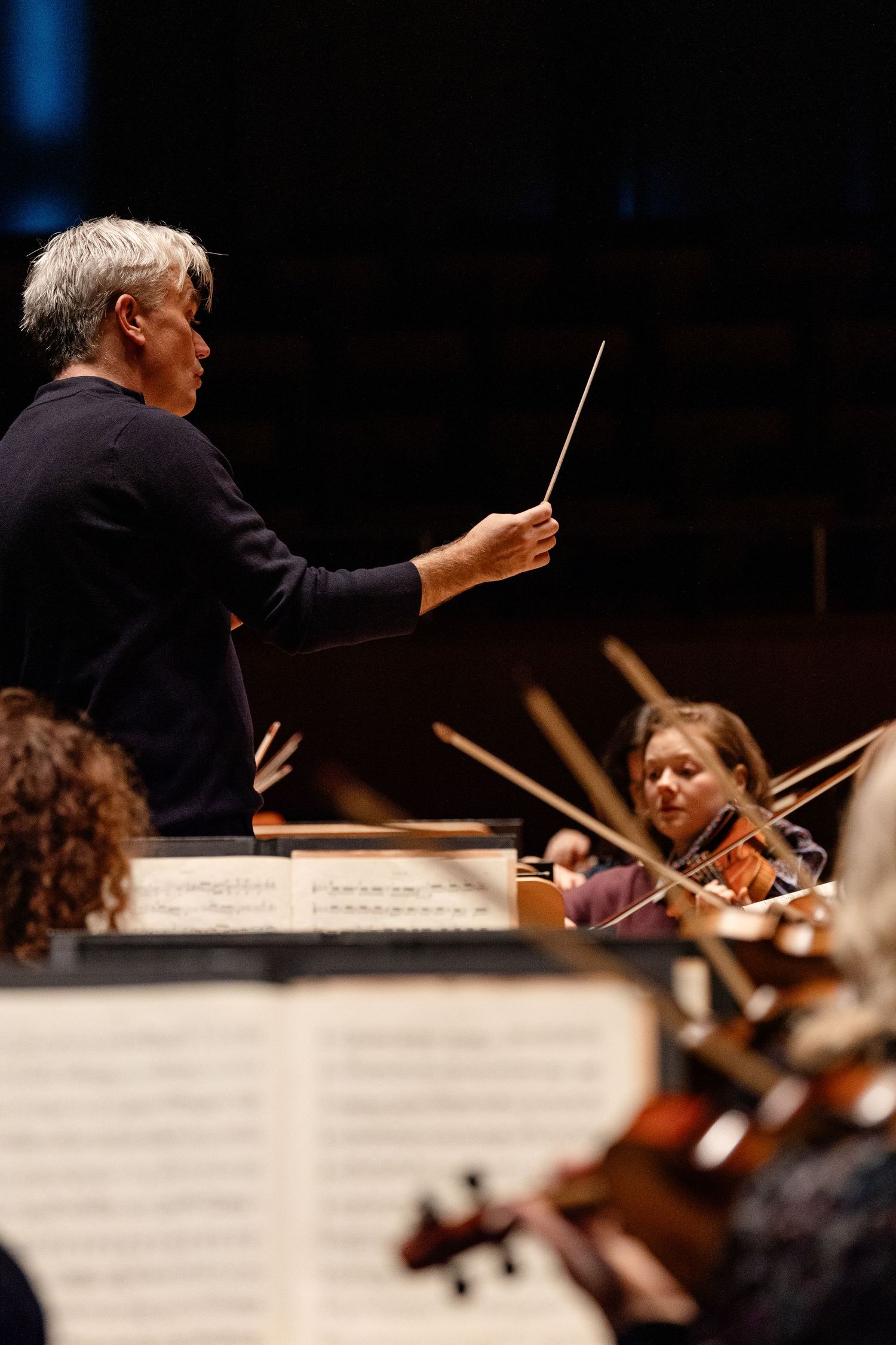 Orchestra conductor leading violinists with baton during a classical music performance in a concert hall.