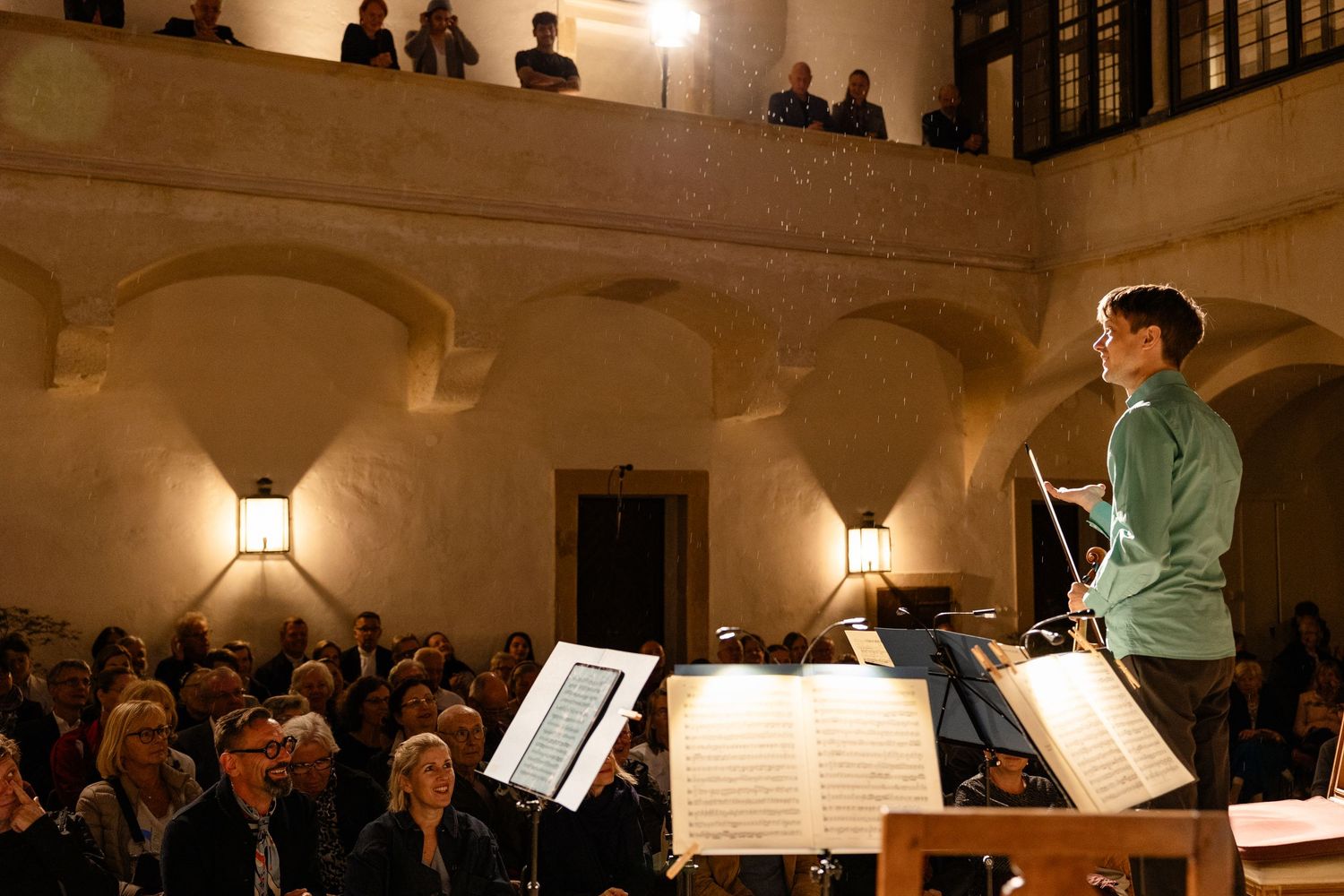 Conductor leading an orchestra performance in an elegant arched concert hall with attentive audience on multiple levels.