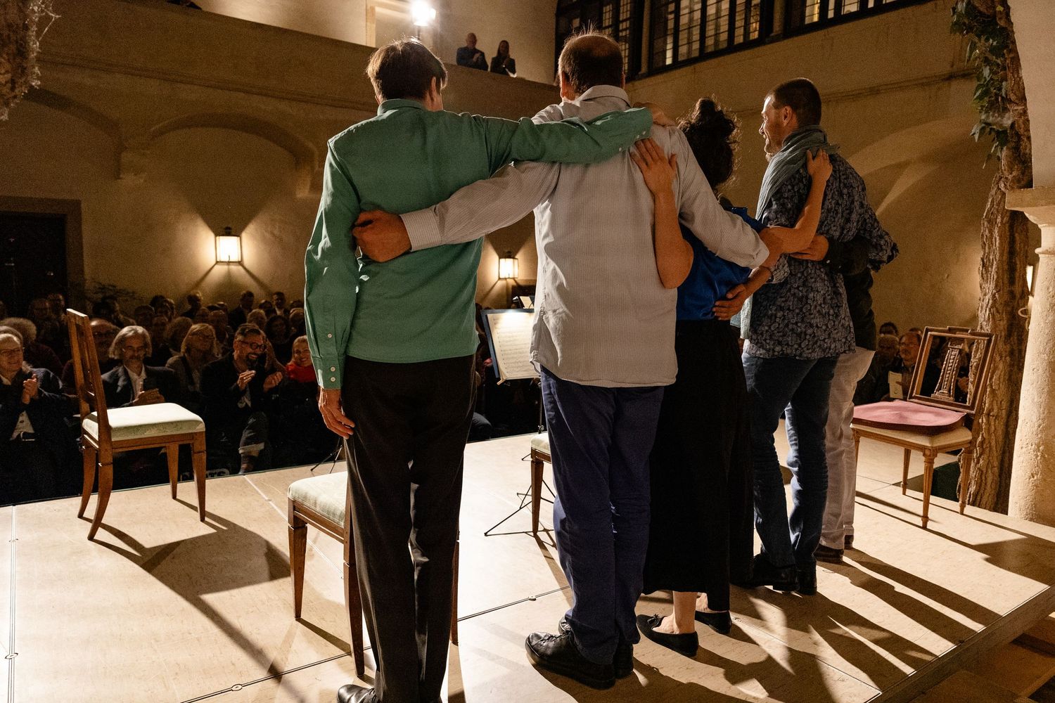 Four people embrace on stage before an audience in an elegant room with bookshelves and warm lighting.