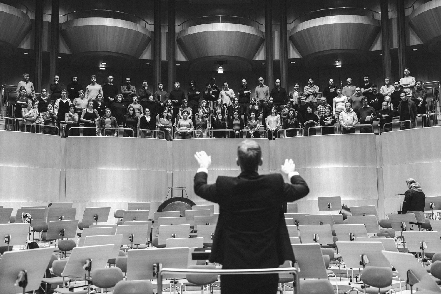 Orchestra conductor leads choir rehearsal in modern concert hall, black and white photography.