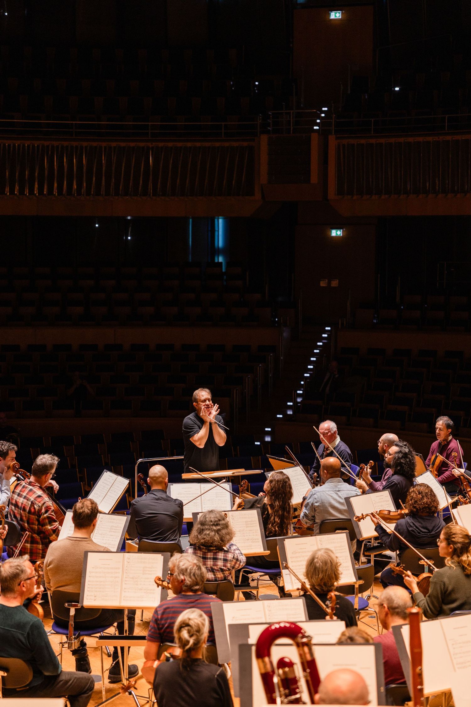 Orchestra rehearsal with conductor leading musicians on stage in a concert hall with empty seats in background.