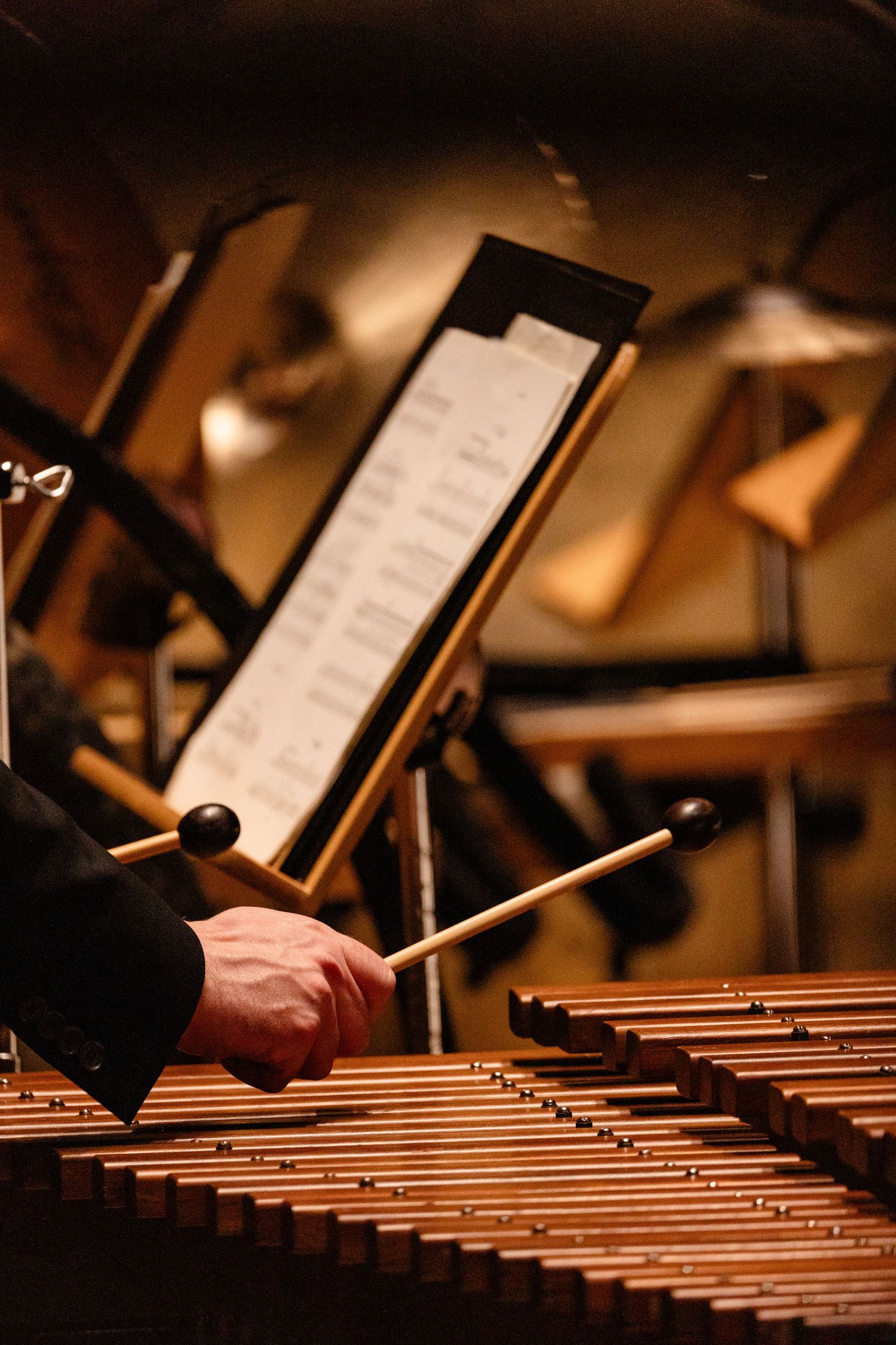 Musician playing marimba with mallets during an orchestra performance, with sheet music on stands in the background.