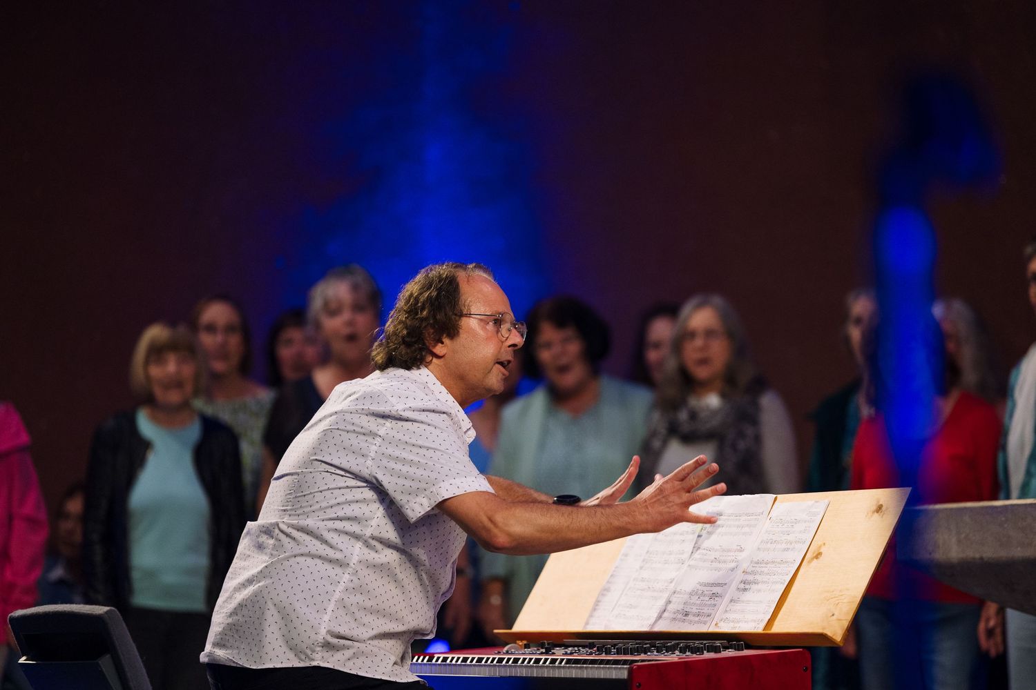 A conductor leads a choir rehearsal, gesturing expressively at a music stand with sheet music indoors.