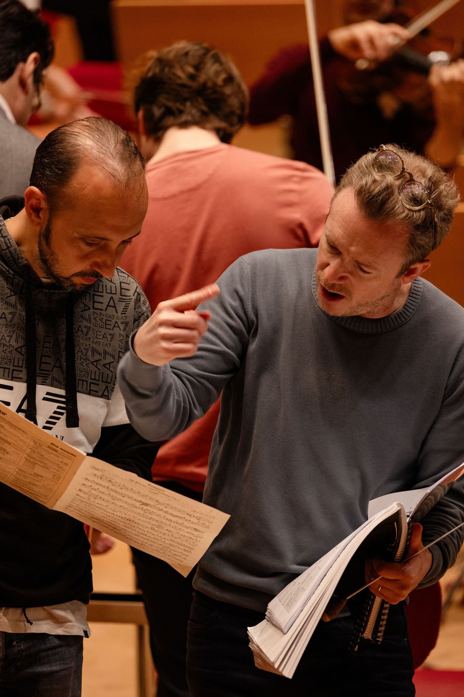 Two musicians review sheet music together during an orchestra rehearsal in a concert hall.