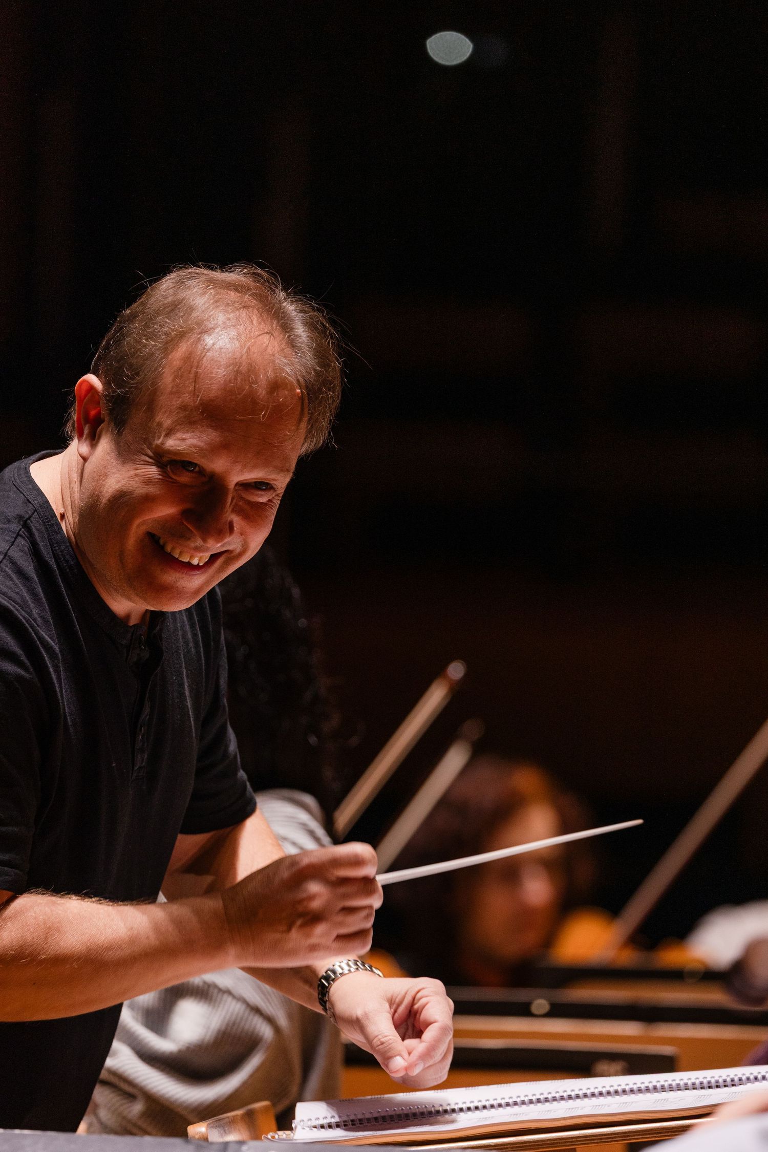 Smiling male orchestra conductor holding a baton while leading a performance on a dimly lit concert stage.