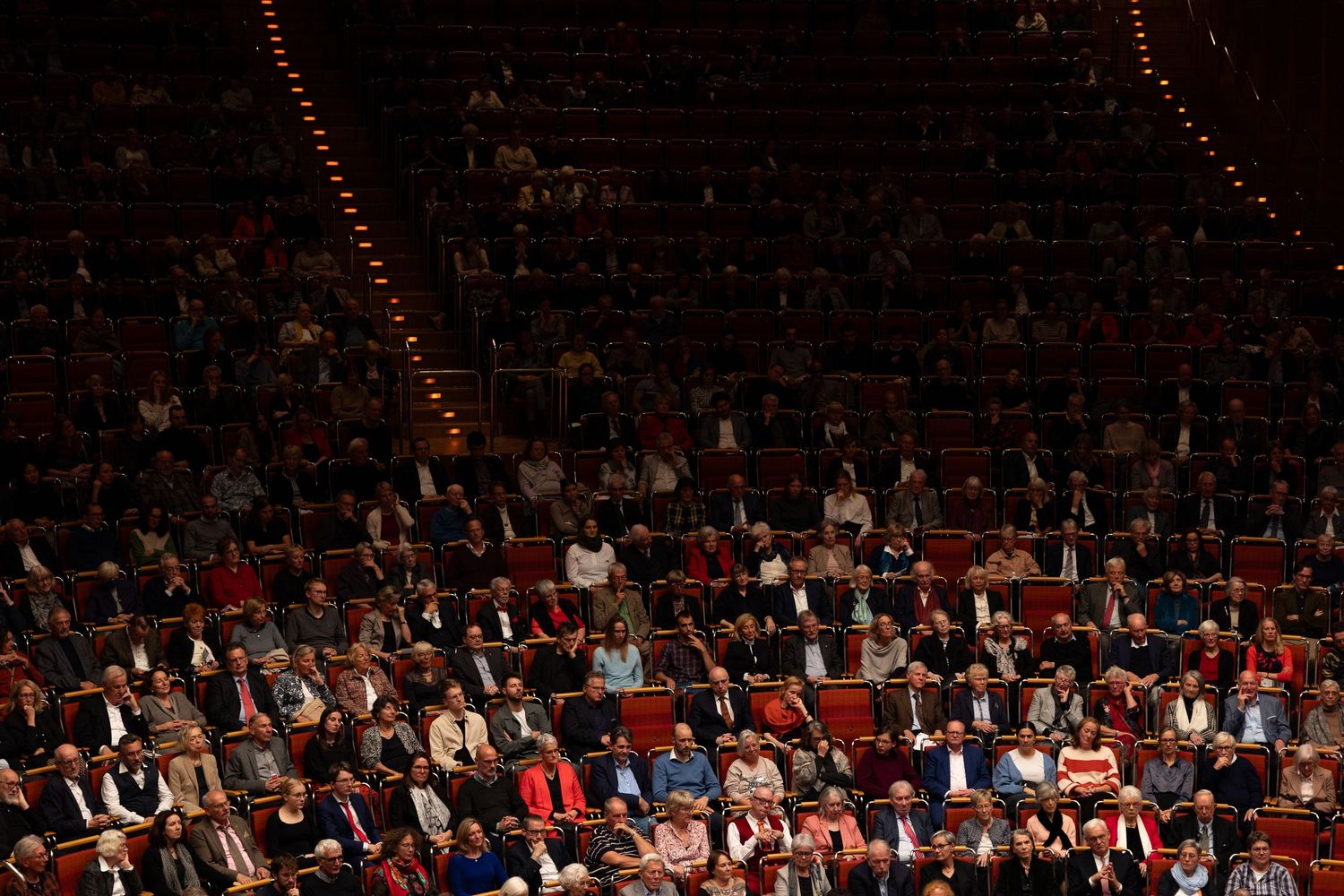 Packed theater audience seated in red chairs across multiple levels during a live performance event.