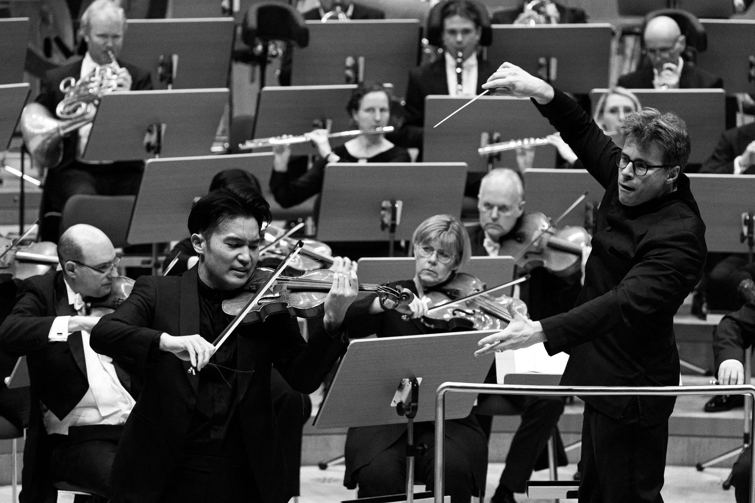 Black and white photo of a violinist performing with an orchestra conductor raising baton during a classical music concert.
