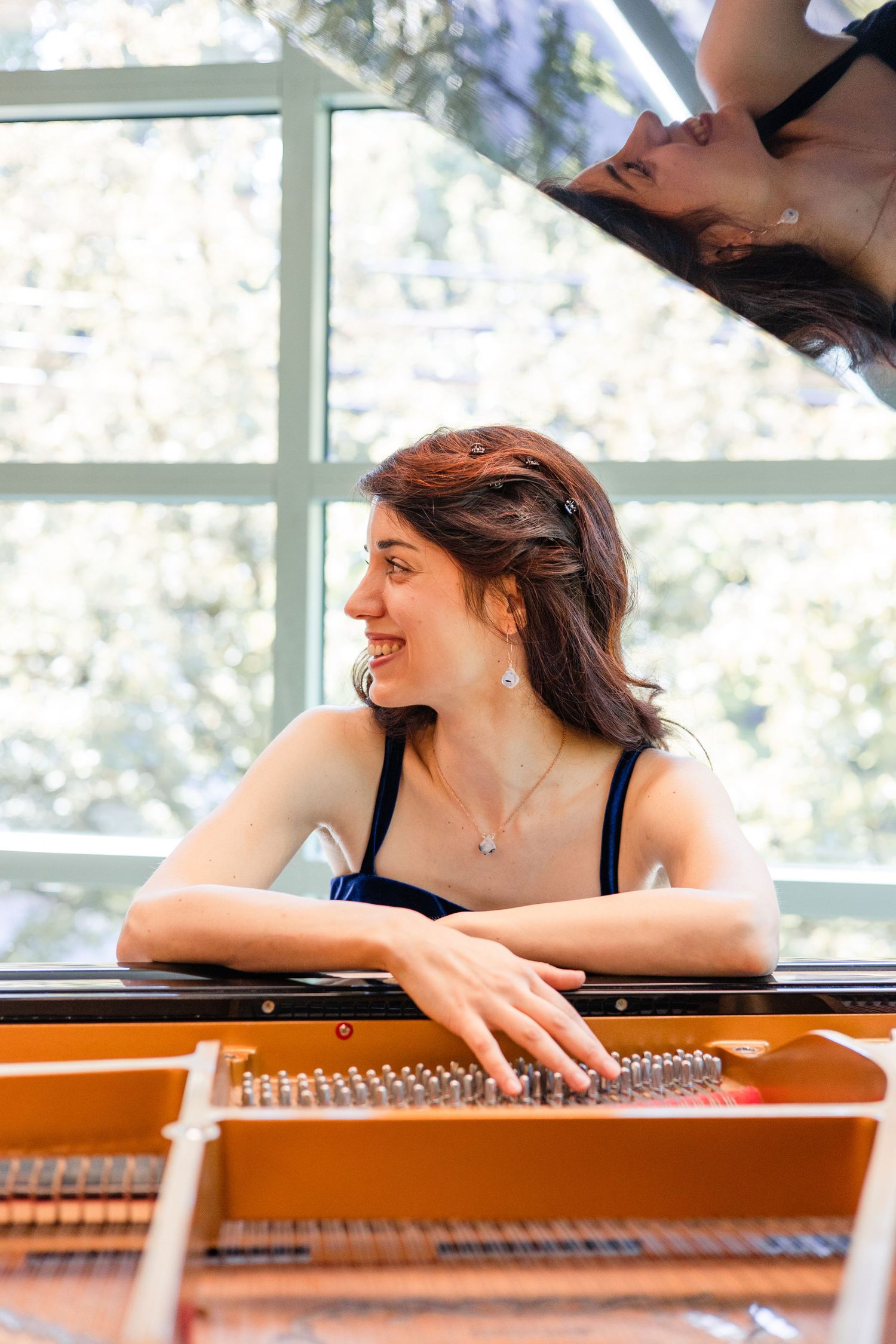 Smiling woman leaning on an open grand piano, with her reflection visible in the raised lid.