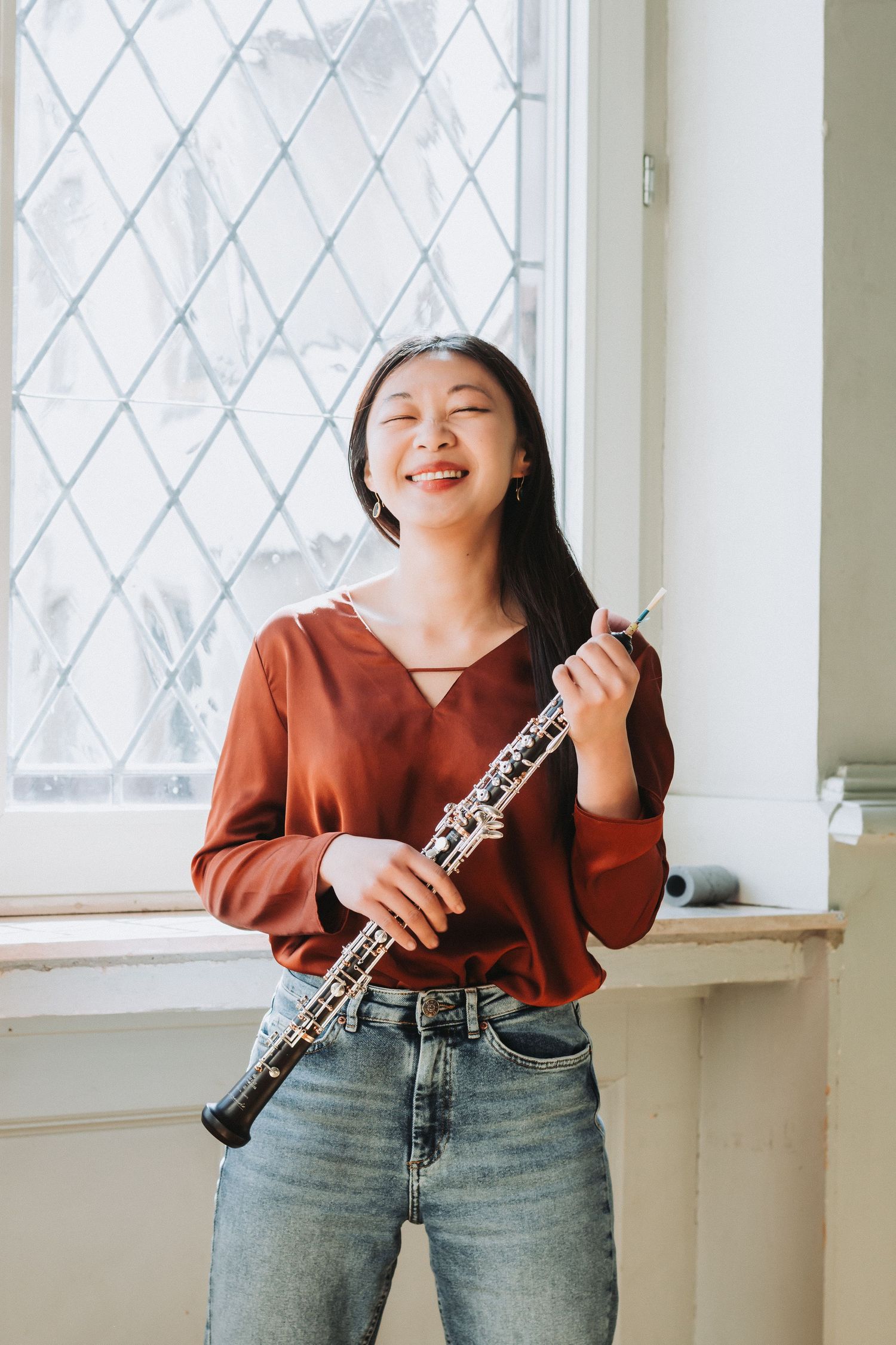 A musician poses playfully with an oboe instrument in front of a bright window wearing a rust-colored sweater and jeans.