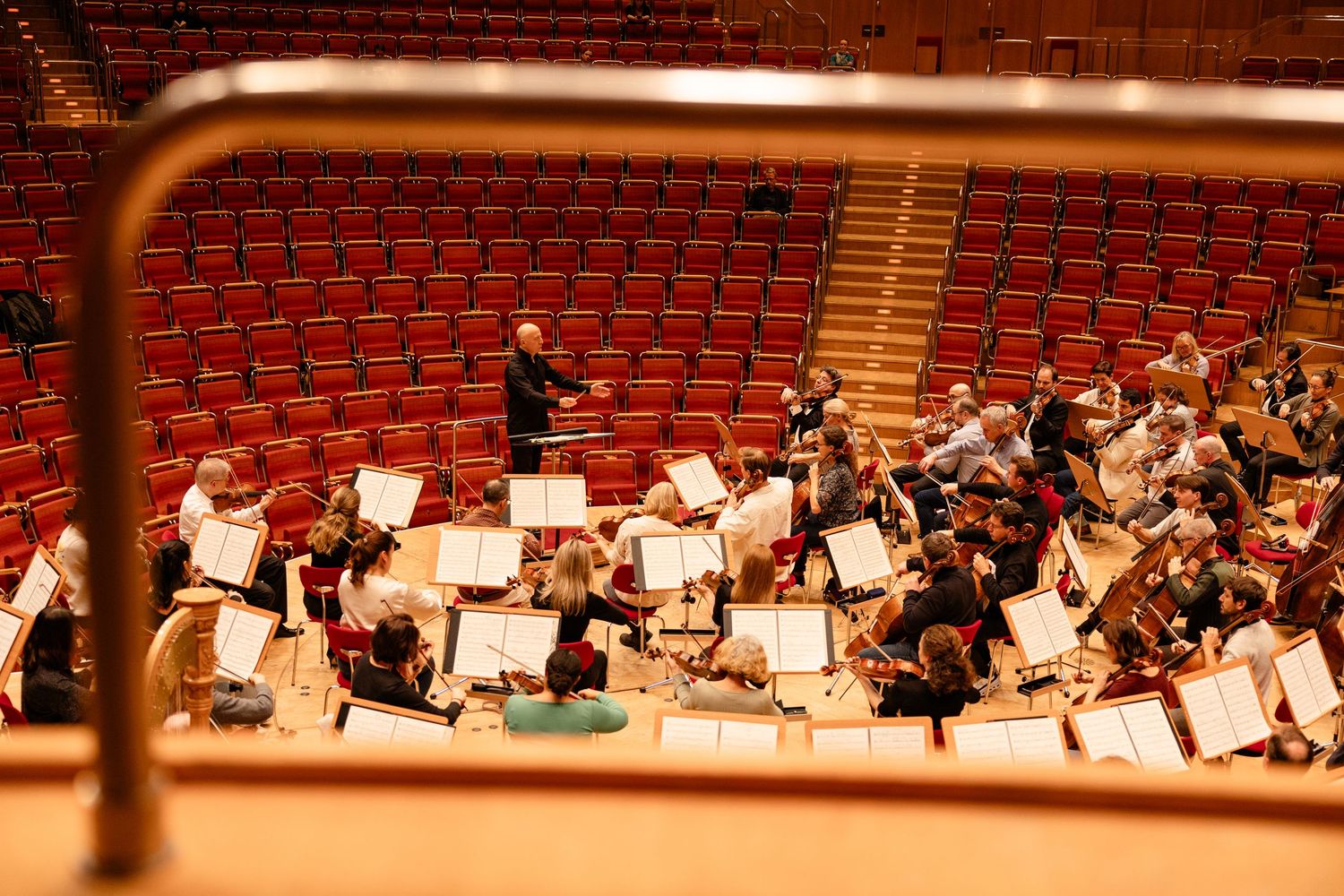 Orchestra rehearsing in empty concert hall, conductor leading musicians with music stands and red seats in background.