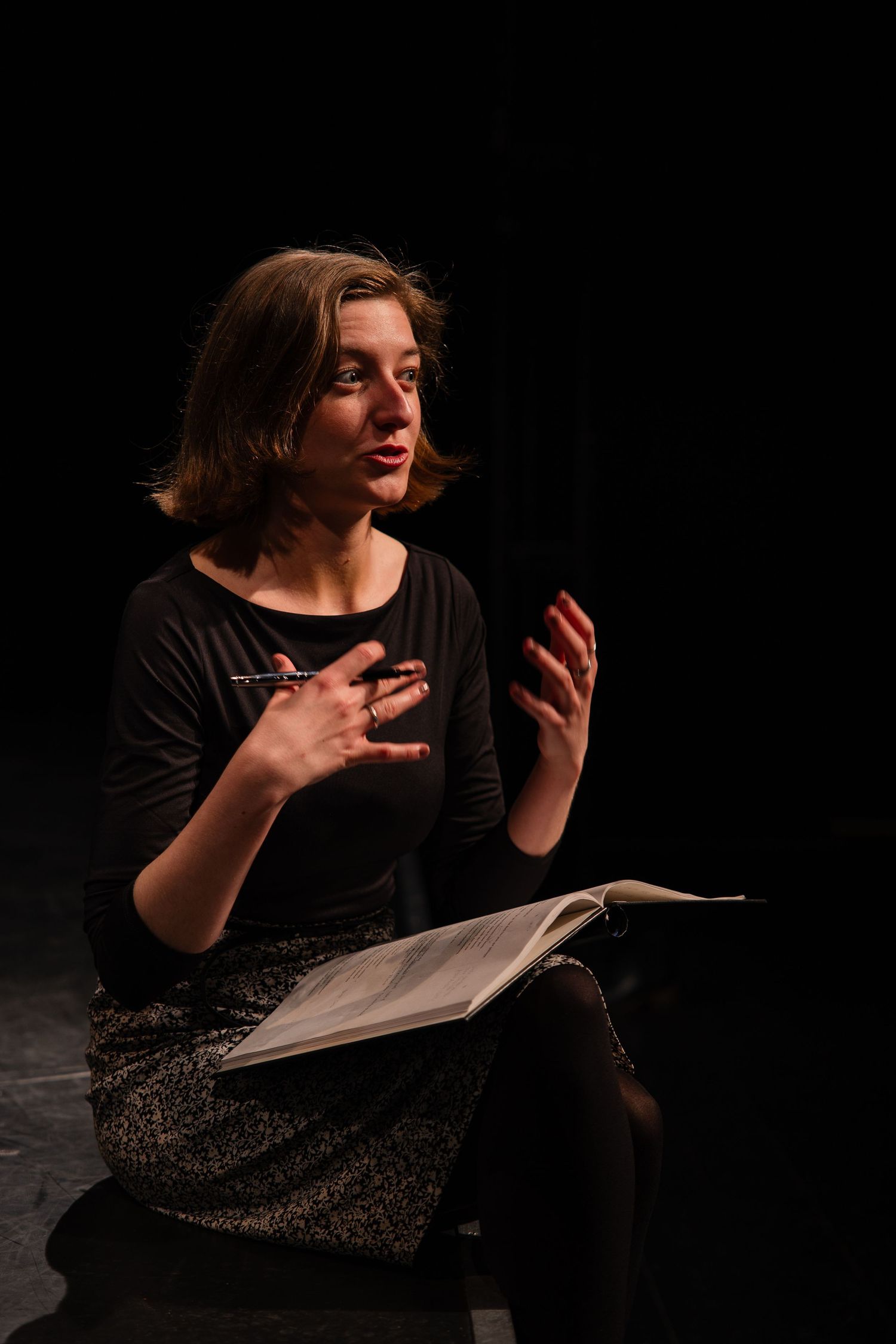 A young woman sits on stage holding an open book, gesturing expressively with her hands during a dramatic performance.
