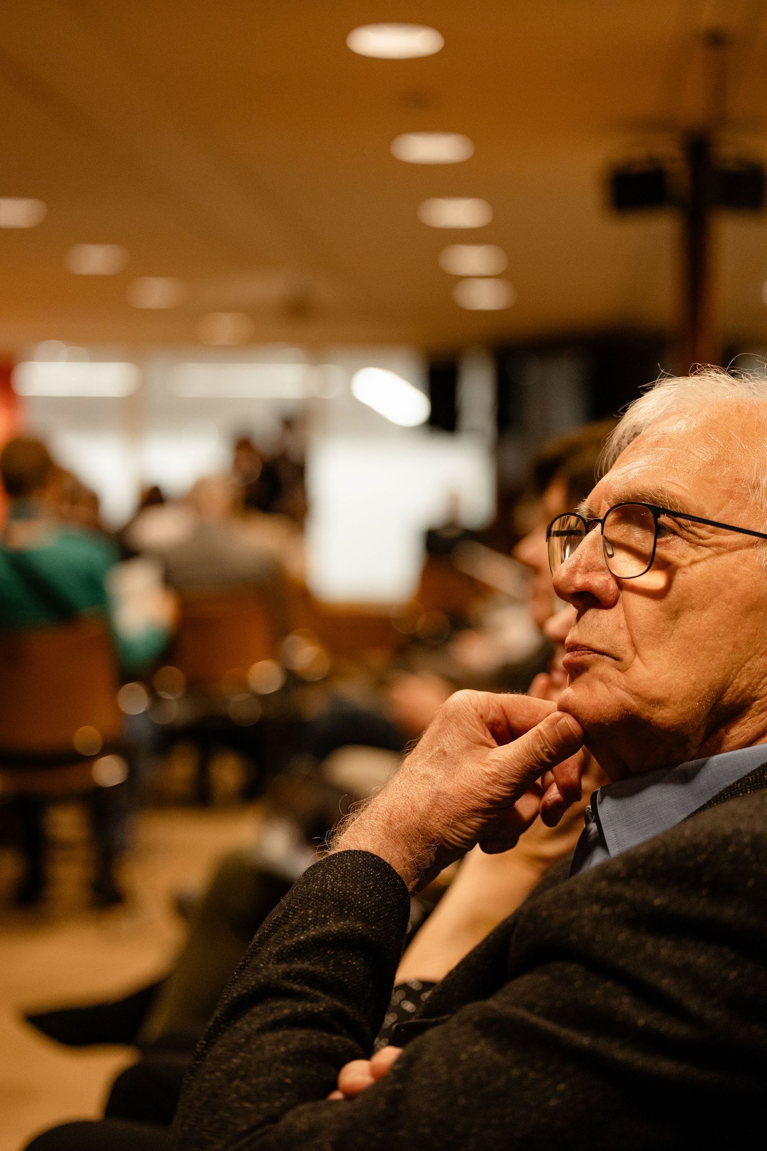 Elderly man with glasses sitting thoughtfully in an audience at a conference, chin resting on hand.