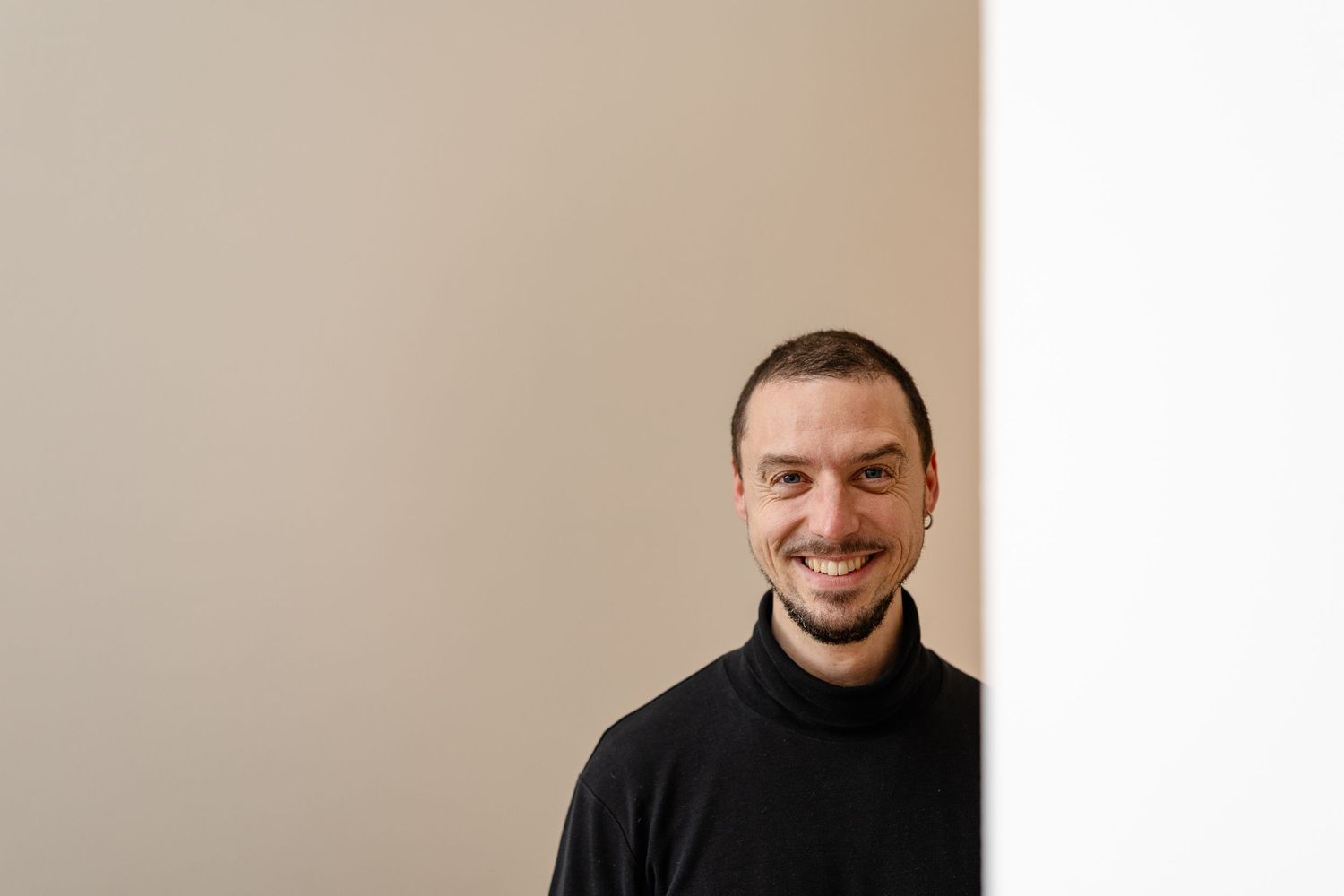 Smiling man with short dark hair wearing a black turtleneck sweater, posing against a neutral beige wall.