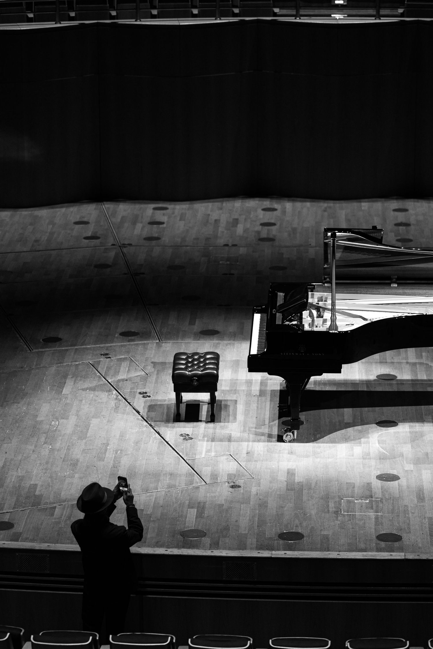 Black and white photo of person photographing a grand piano on an empty concert hall stage.