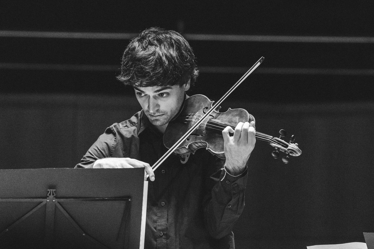 Young male violinist playing violin while reading sheet music, captured in black and white photography.