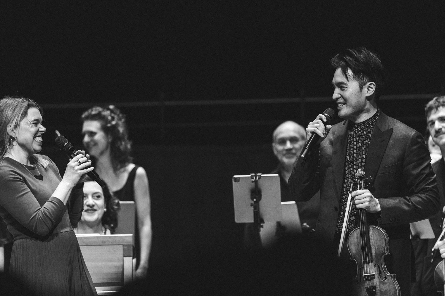 Two performers share a microphone on stage while holding a violin during a black and white concert event.