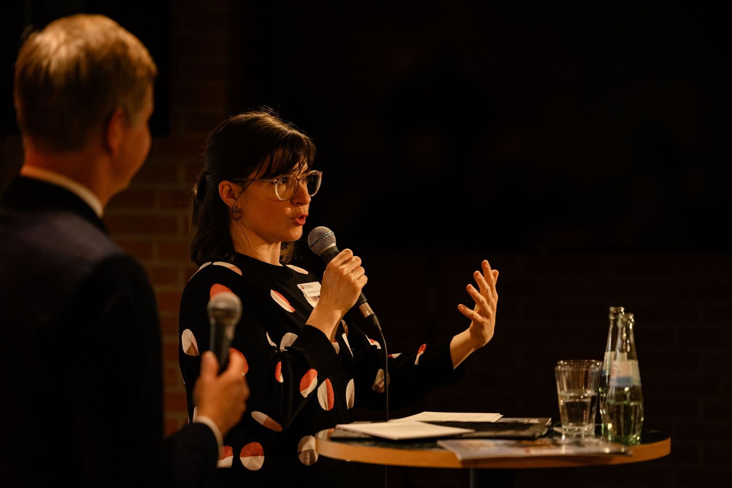 Woman in polka dot dress speaks into microphone at panel discussion event, gesturing expressively on dimly lit stage.