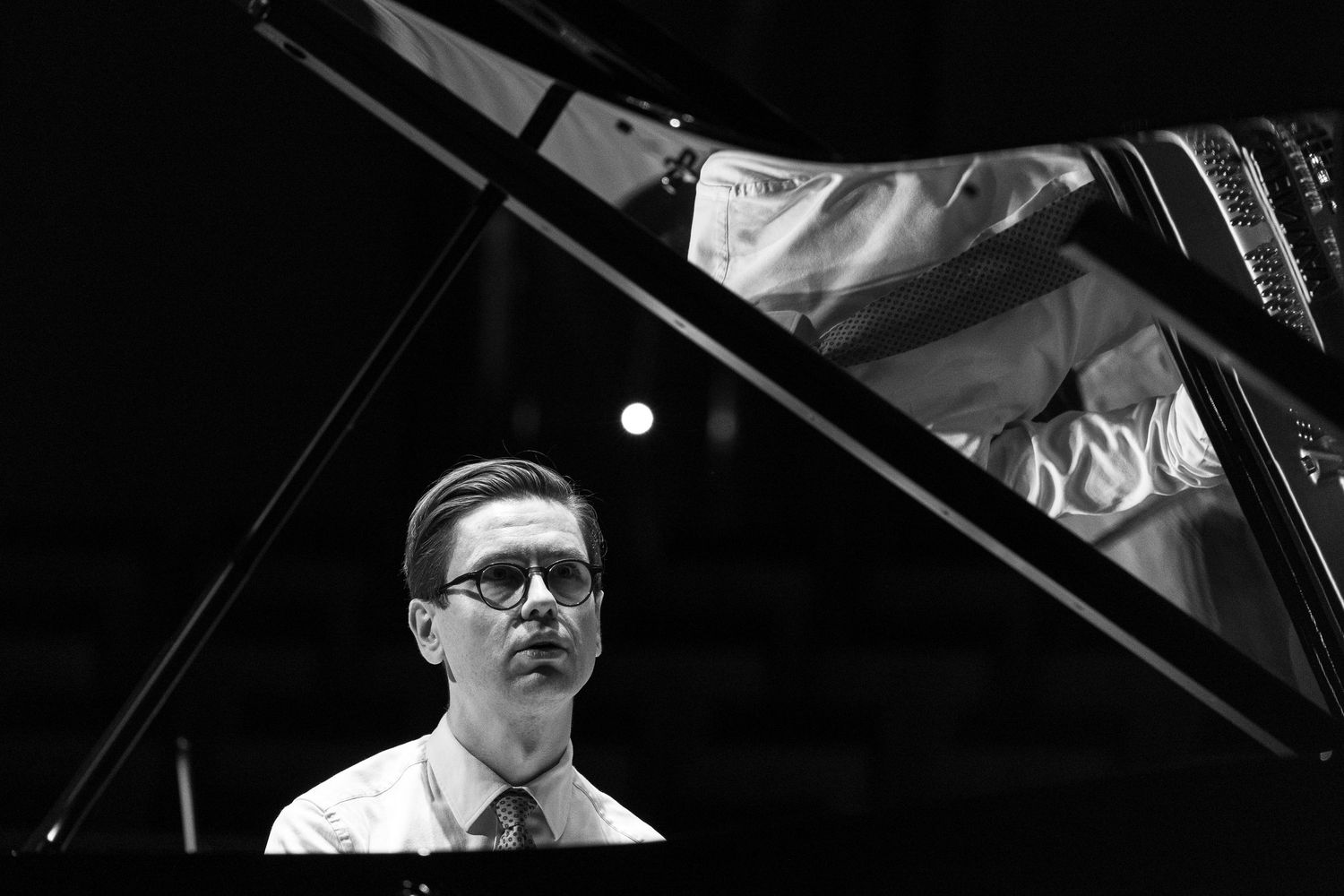 Black and white photo of a male pianist wearing glasses performing at a grand piano on stage.