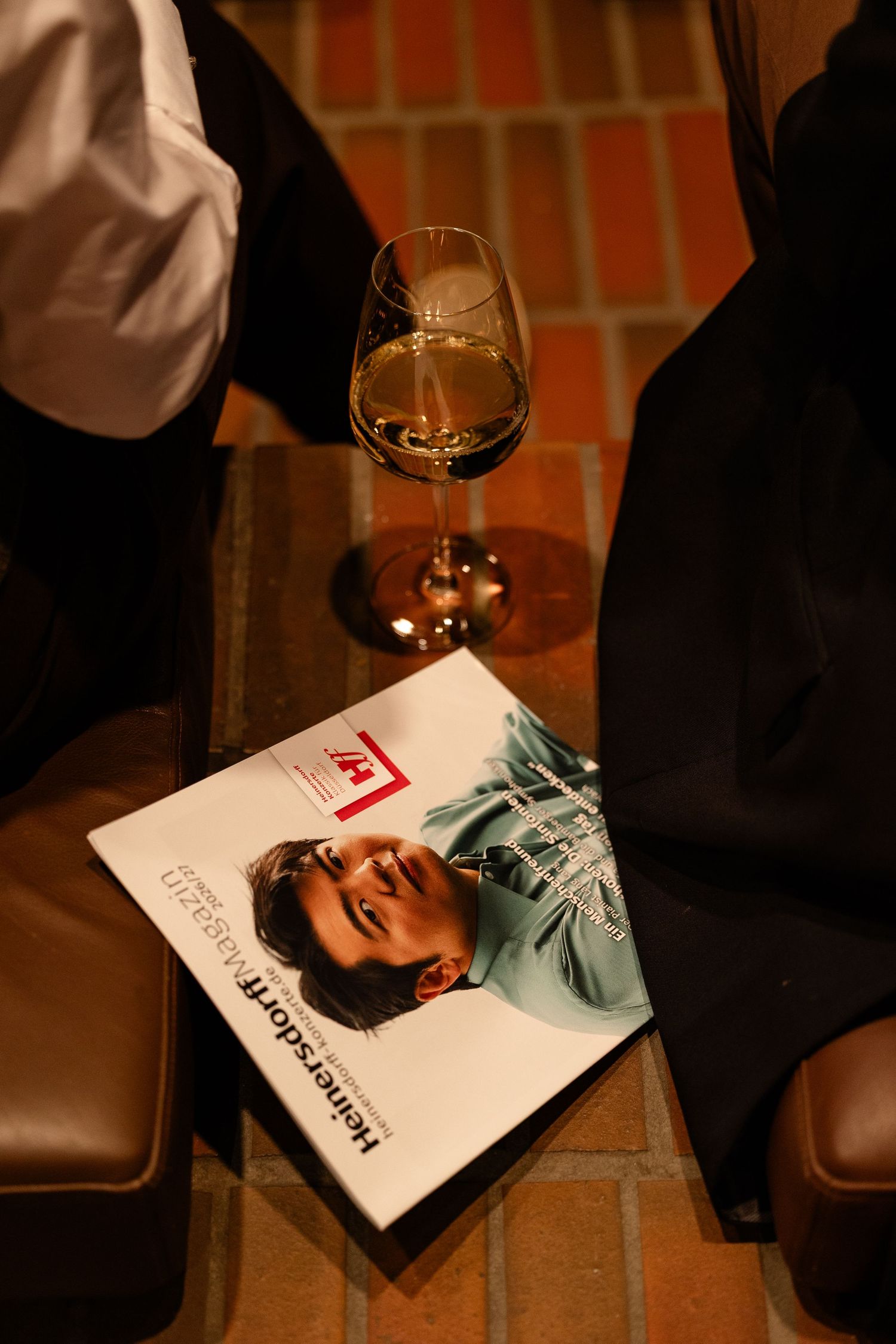 A wine glass with white wine sits on a ledge beside a magazine featuring a man's portrait at an evening event.