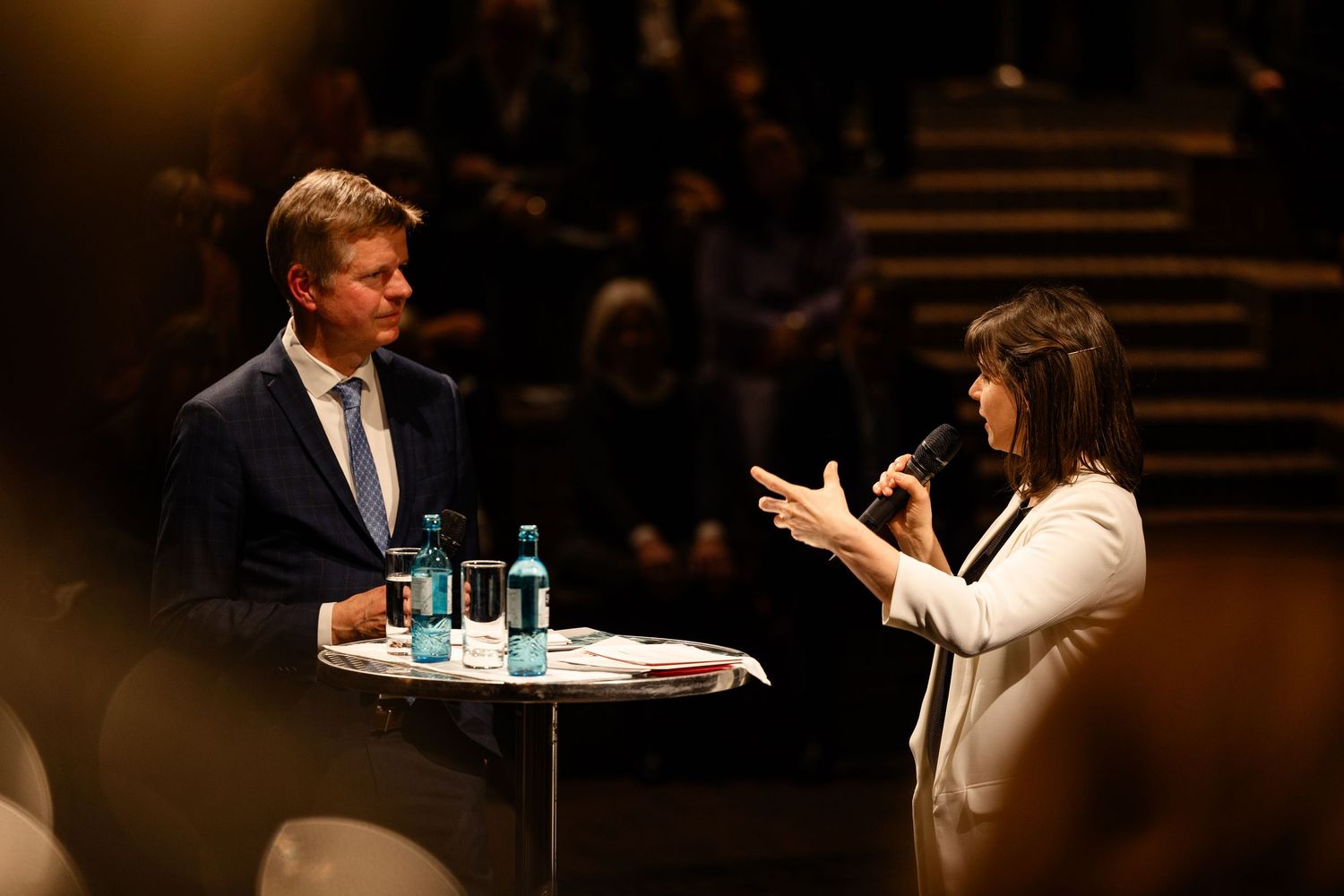 A speaker gestures while seated at a table during a darkly lit presentation or panel discussion.
