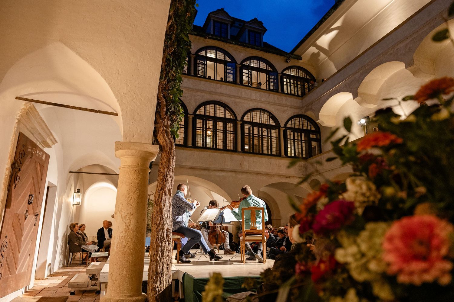 Musicians perform in an elegant courtyard of a historic building at dusk, surrounded by arched windows and flowers.